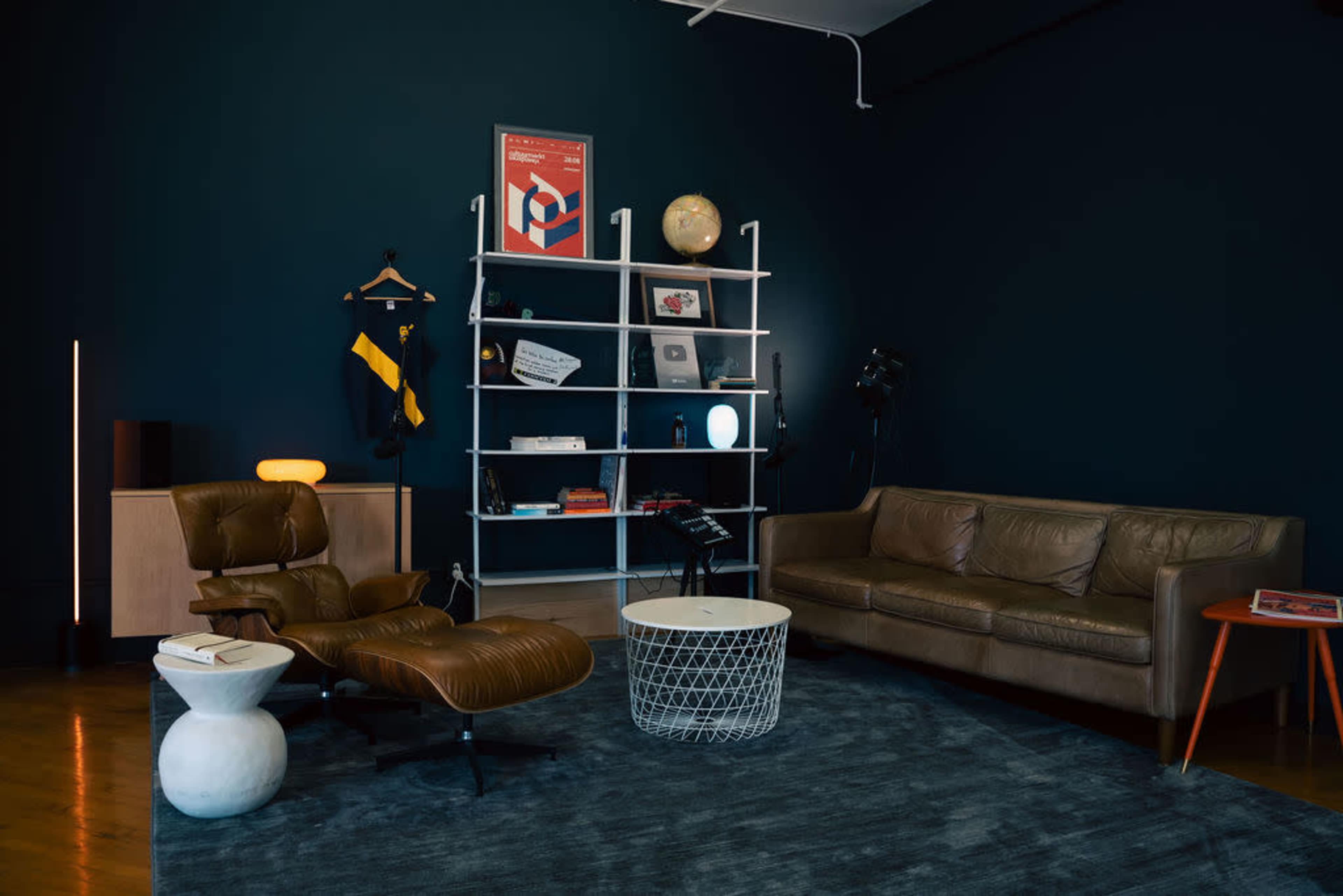 The image shows a modern living room featuring a brown leather sofa, an armchair, a round white coffee table, a bookshelf with various items, and a dark blue wall.
