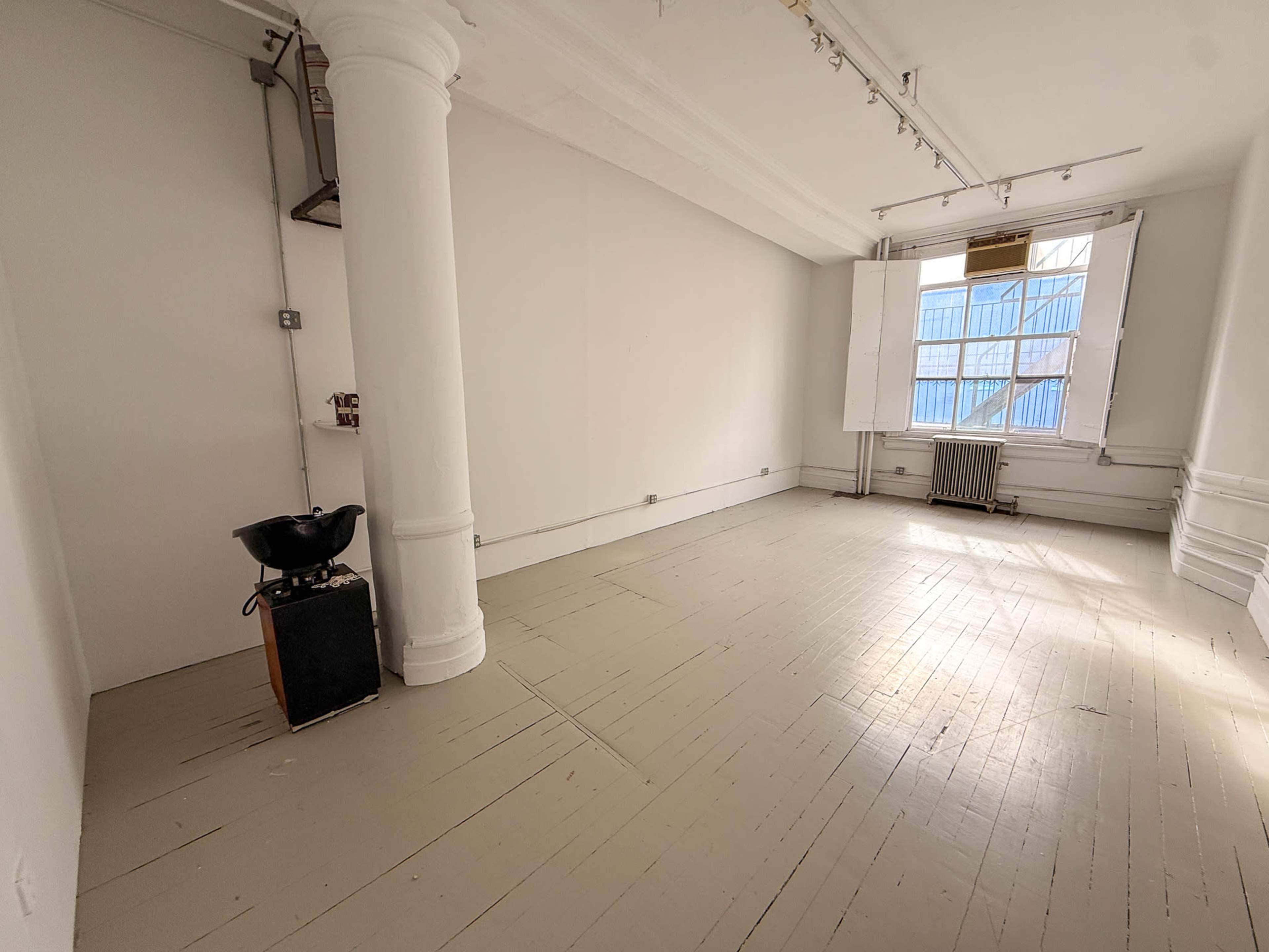 The image shows an empty room with white walls, wooden flooring, a column, and a window partially covered by blue scaffolding.