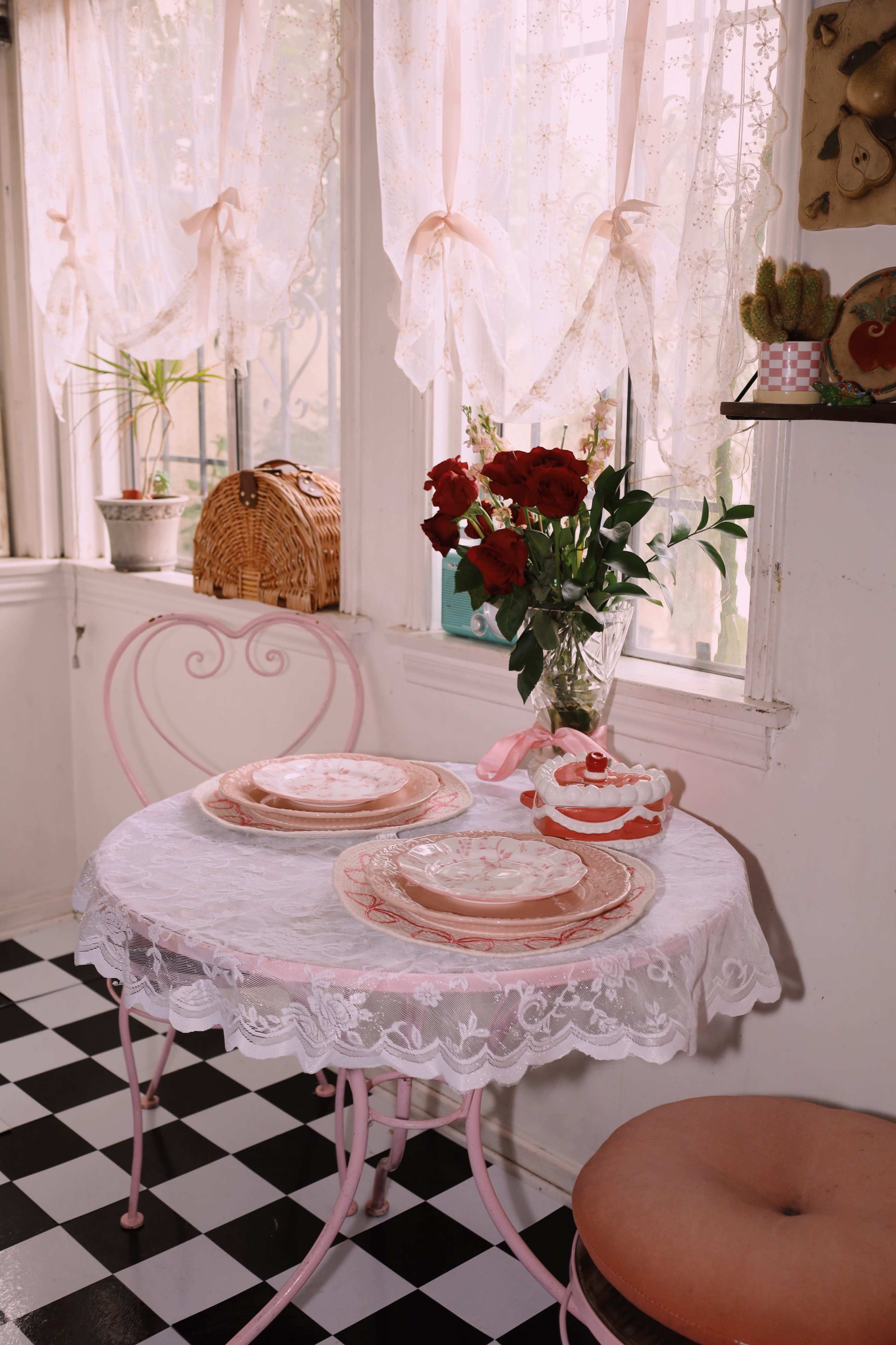 A small dining table with pink chairs is set with plates and a vase of red roses, all in a sunlit corner adorned with sheer curtains.