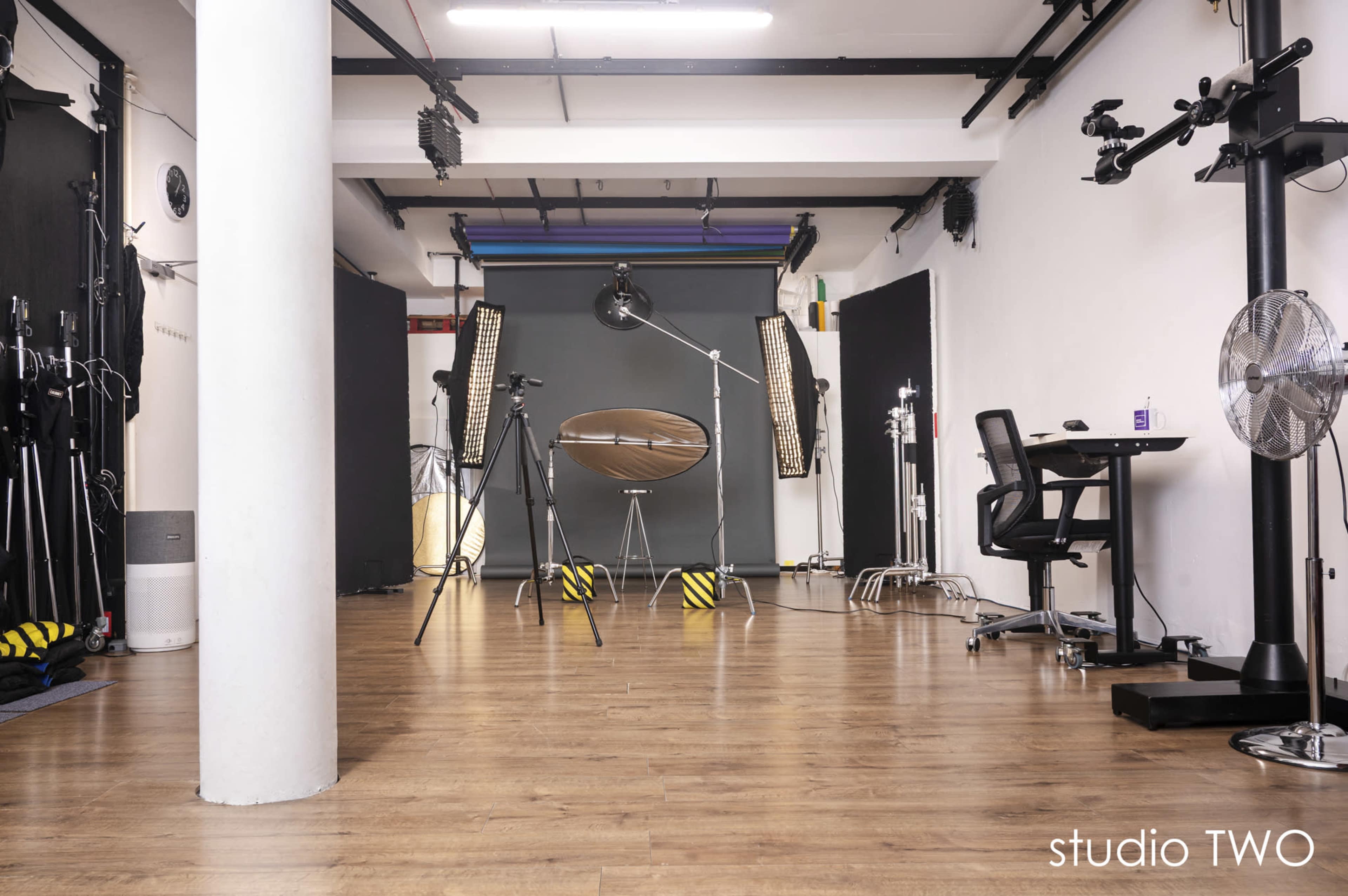 The image shows a photography studio with equipment such as lights, a large reflector, and a backdrop, all arranged in an organized space with wooden flooring.