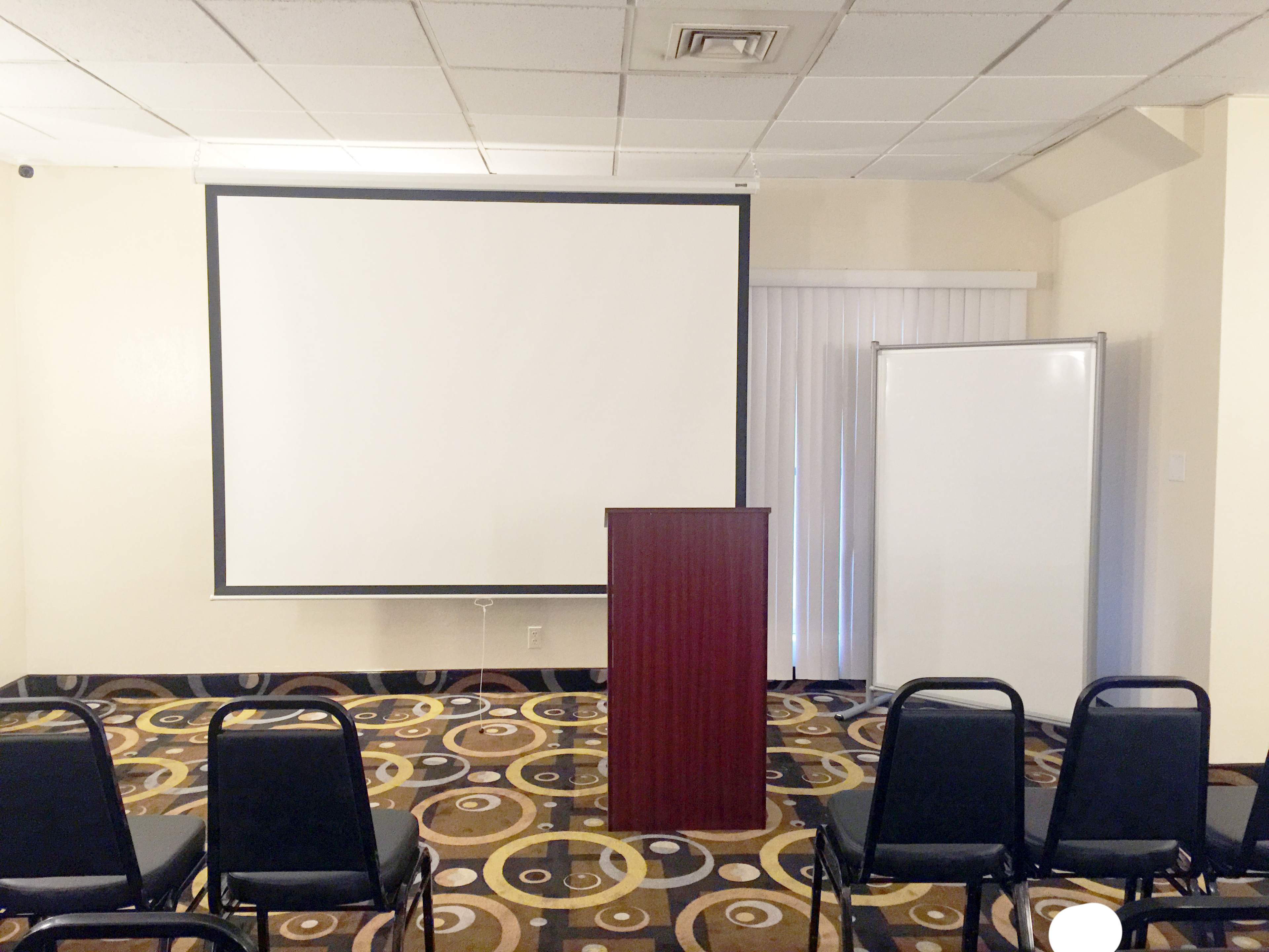 A room is set up for a presentation, featuring a podium, a blank projection screen, and rows of empty chairs.