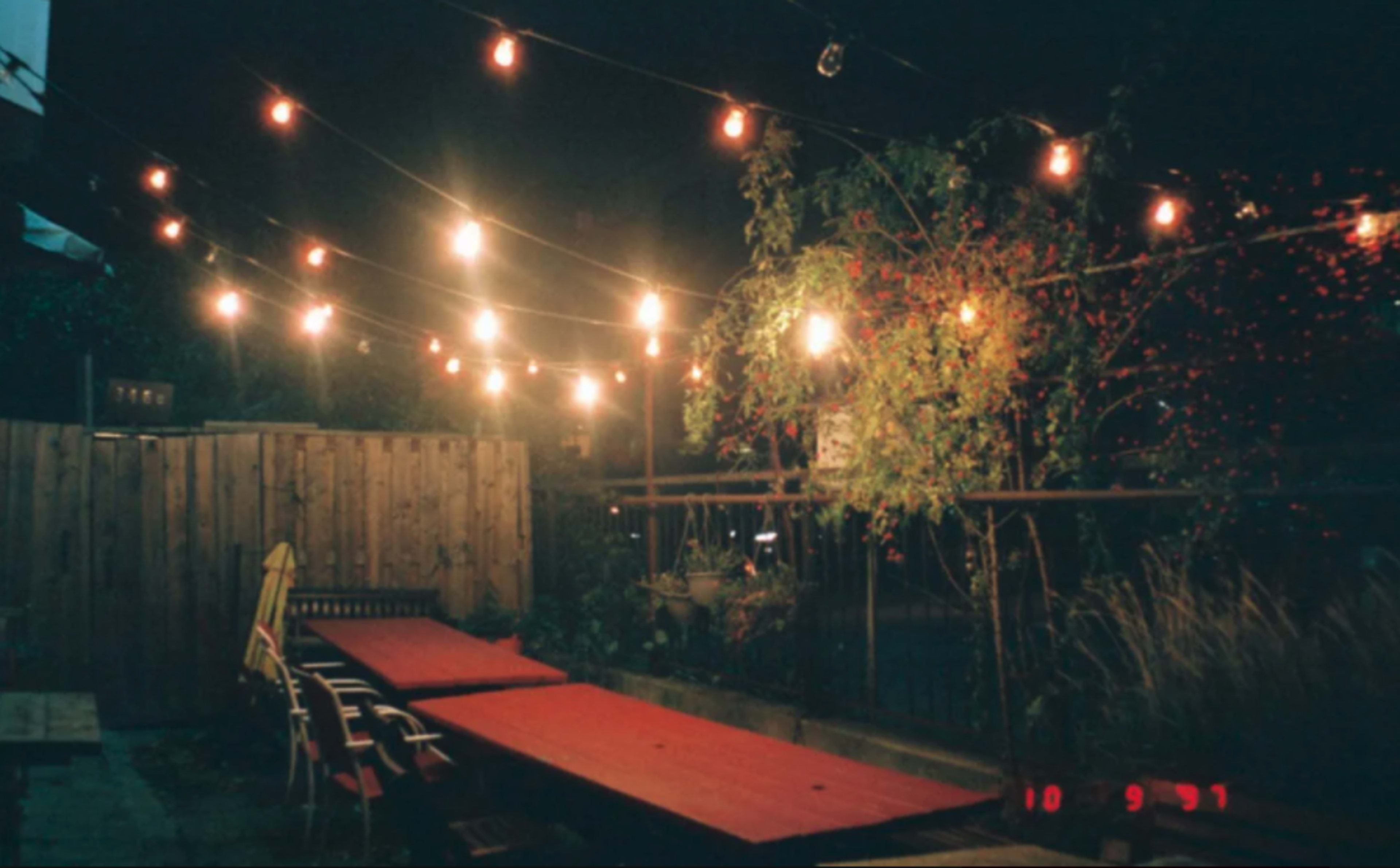 A backyard patio at night, illuminated by string lights above a long red table and wooden chairs.