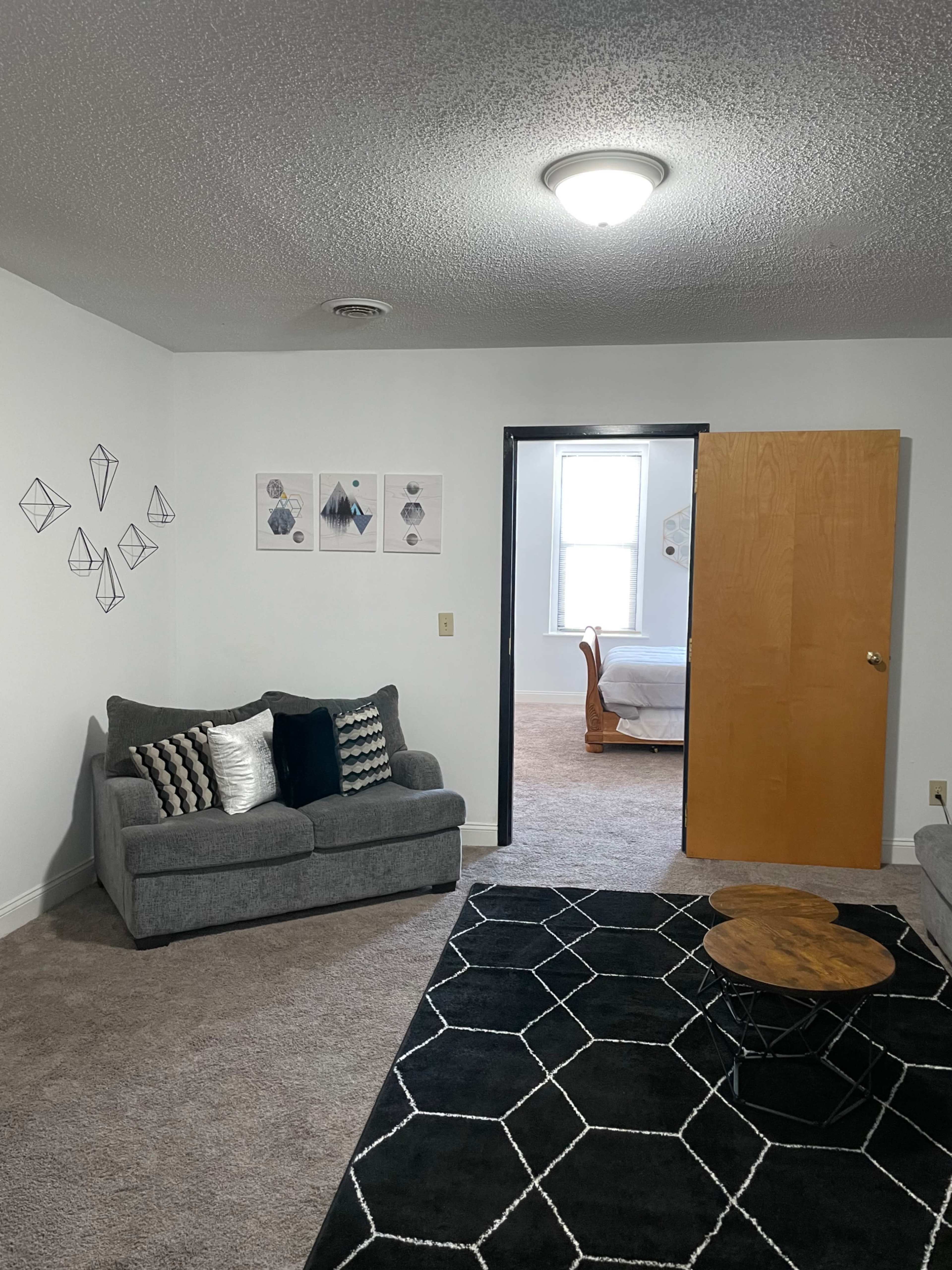 A living area with a gray sofa, a round wooden coffee table, and a doorway leading to a bedroom in a carpeted room.