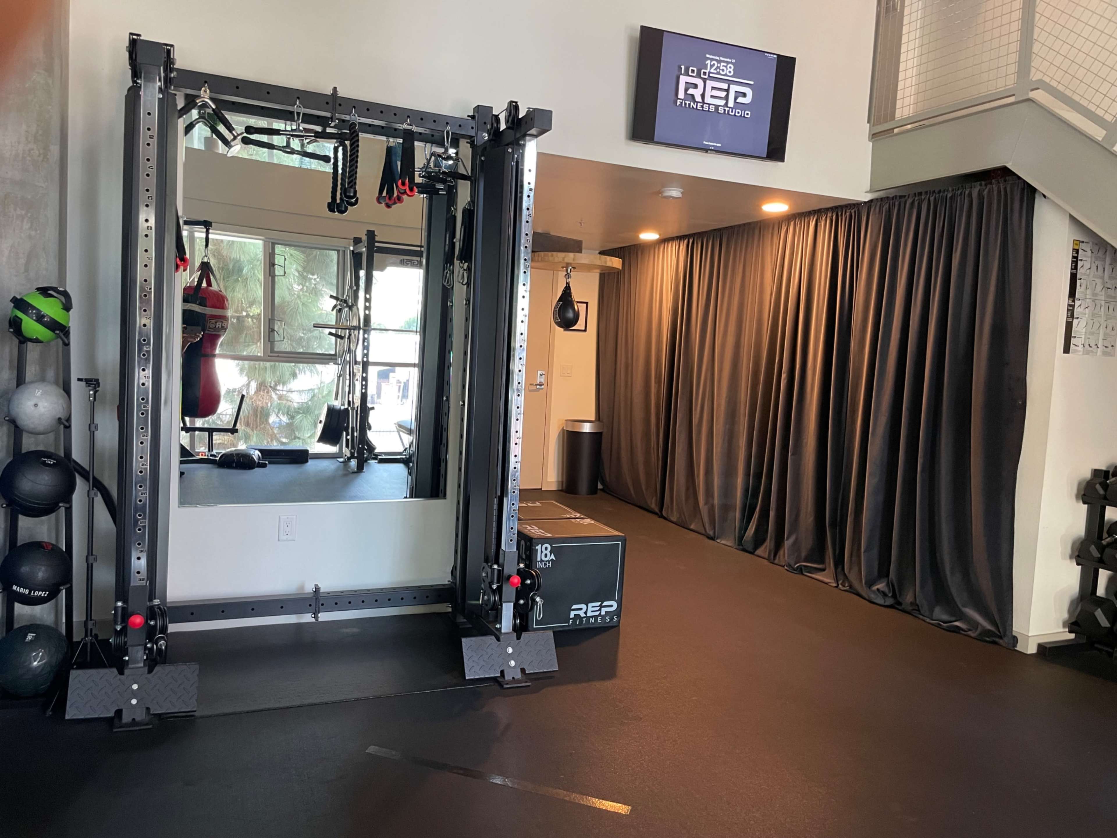 The image shows a home gym with a mirror, a weightlifting rack, a punching bag, and a curtained area on one side.
