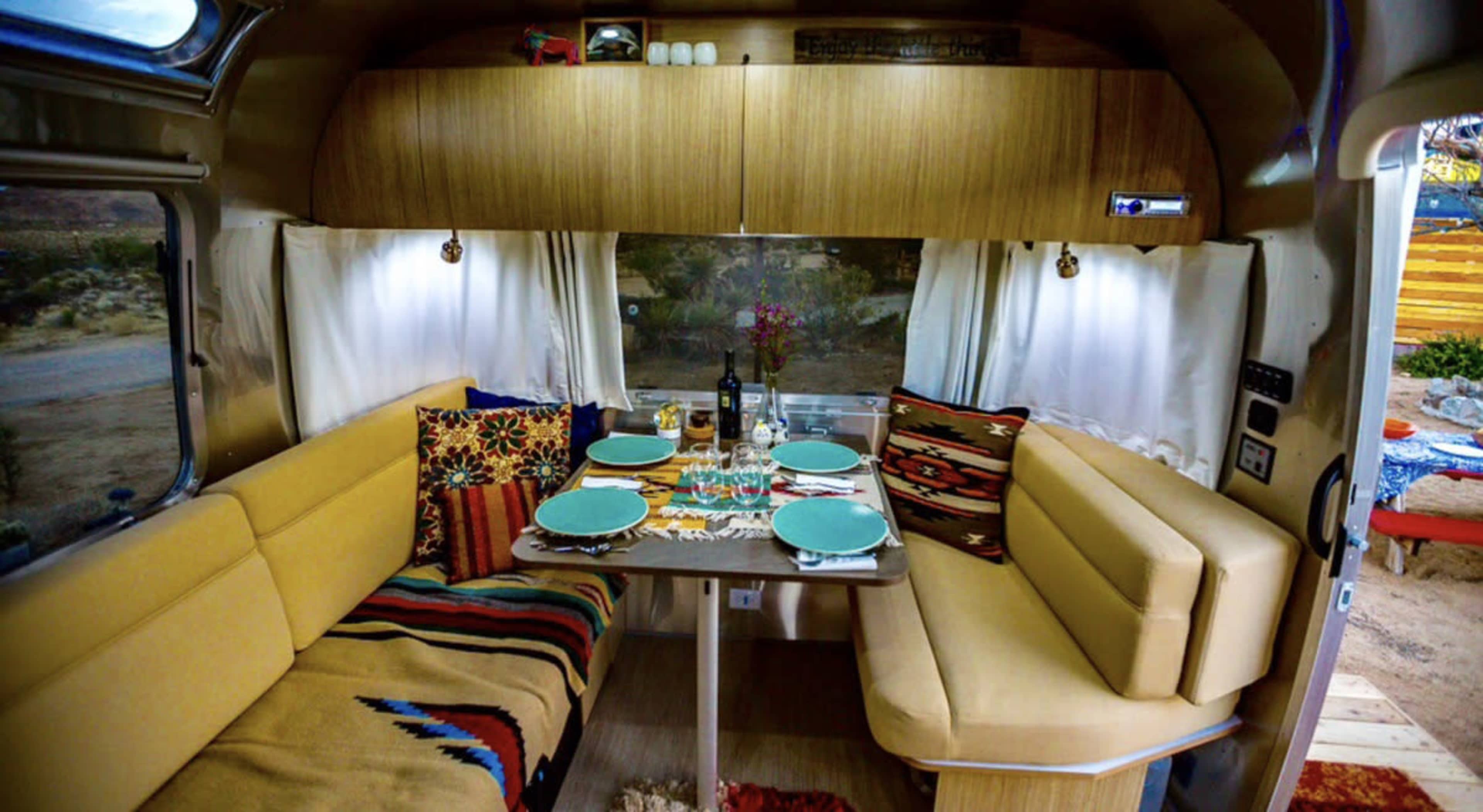 The image shows a cozy dining area inside a retro-style trailer, featuring a table set for a meal with colorful plates and decorative cushions.