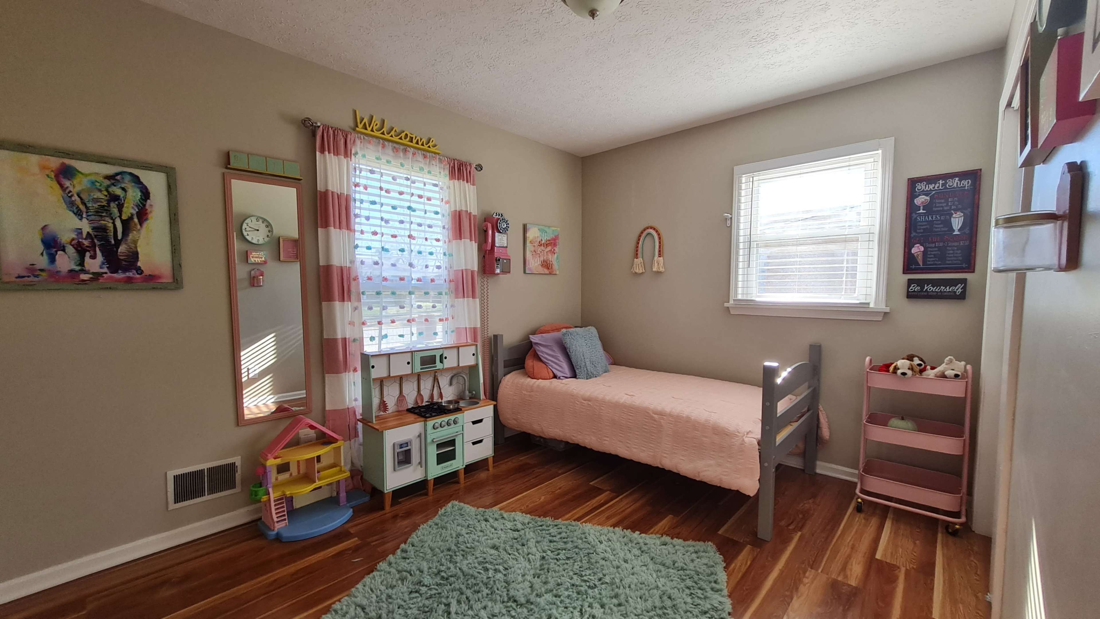 A child's bedroom with a pink bed, a small playhouse, a green rug, and windows adorned with light curtains.