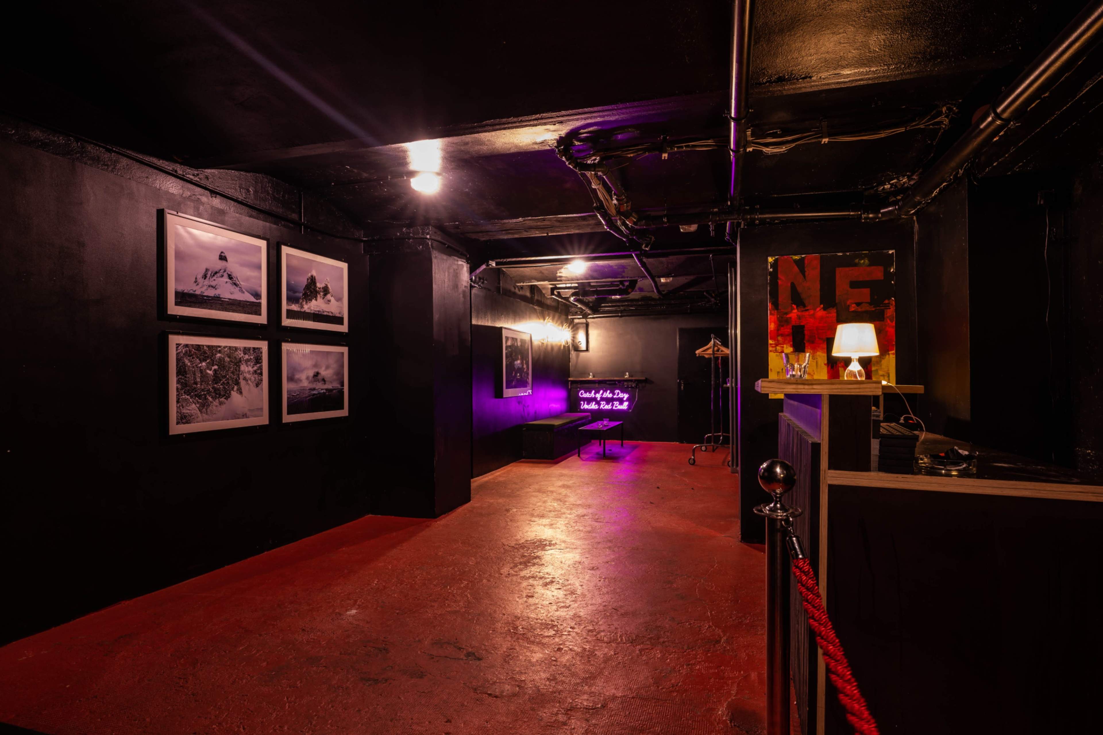 The image depicts a dimly lit hallway with black walls, featuring framed photographs on one side and a reception area with a lamp and red rope barrier at the entrance.