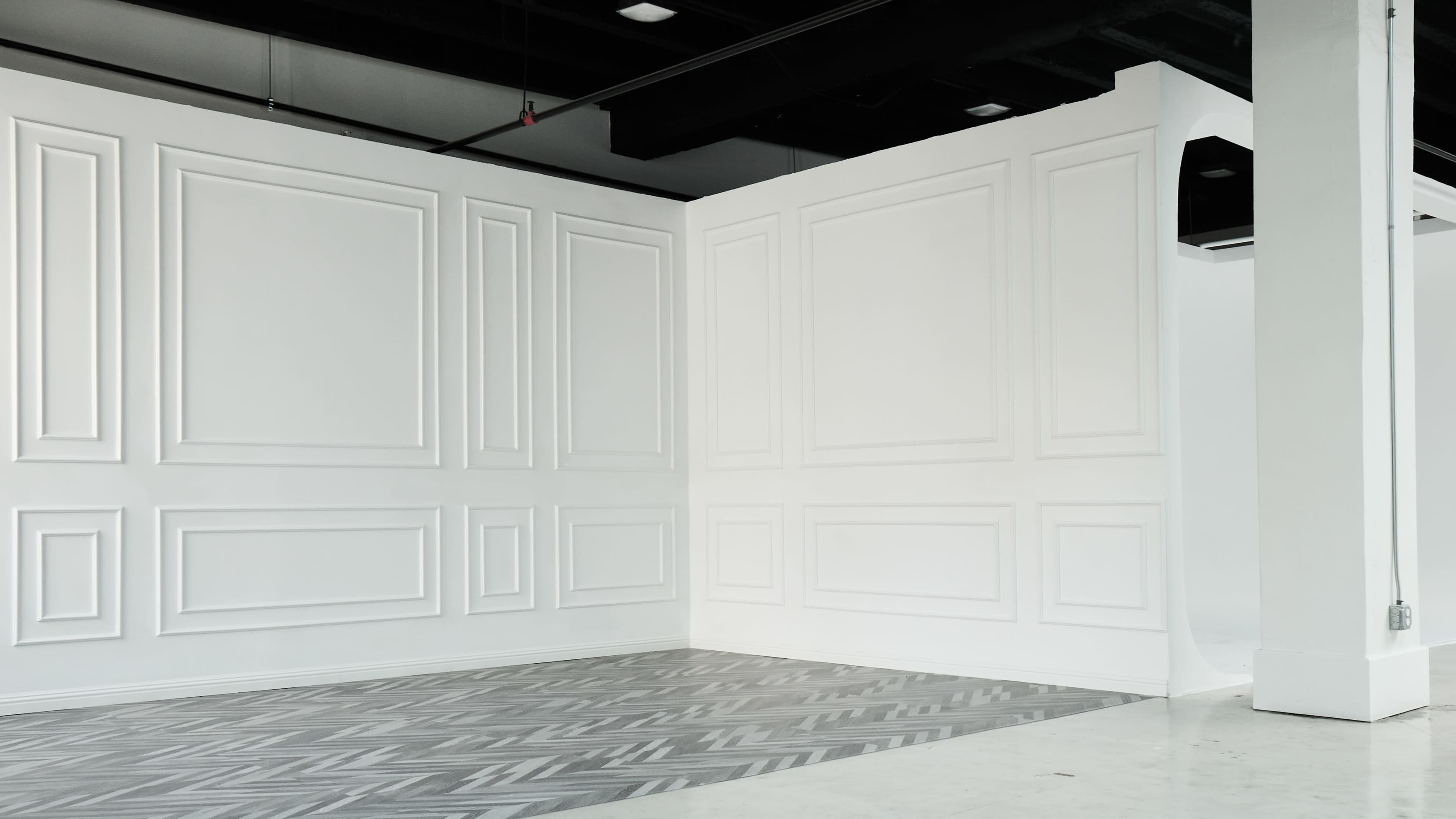 A spacious, empty room with white walls featuring decorative paneling and a herringbone-patterned floor.