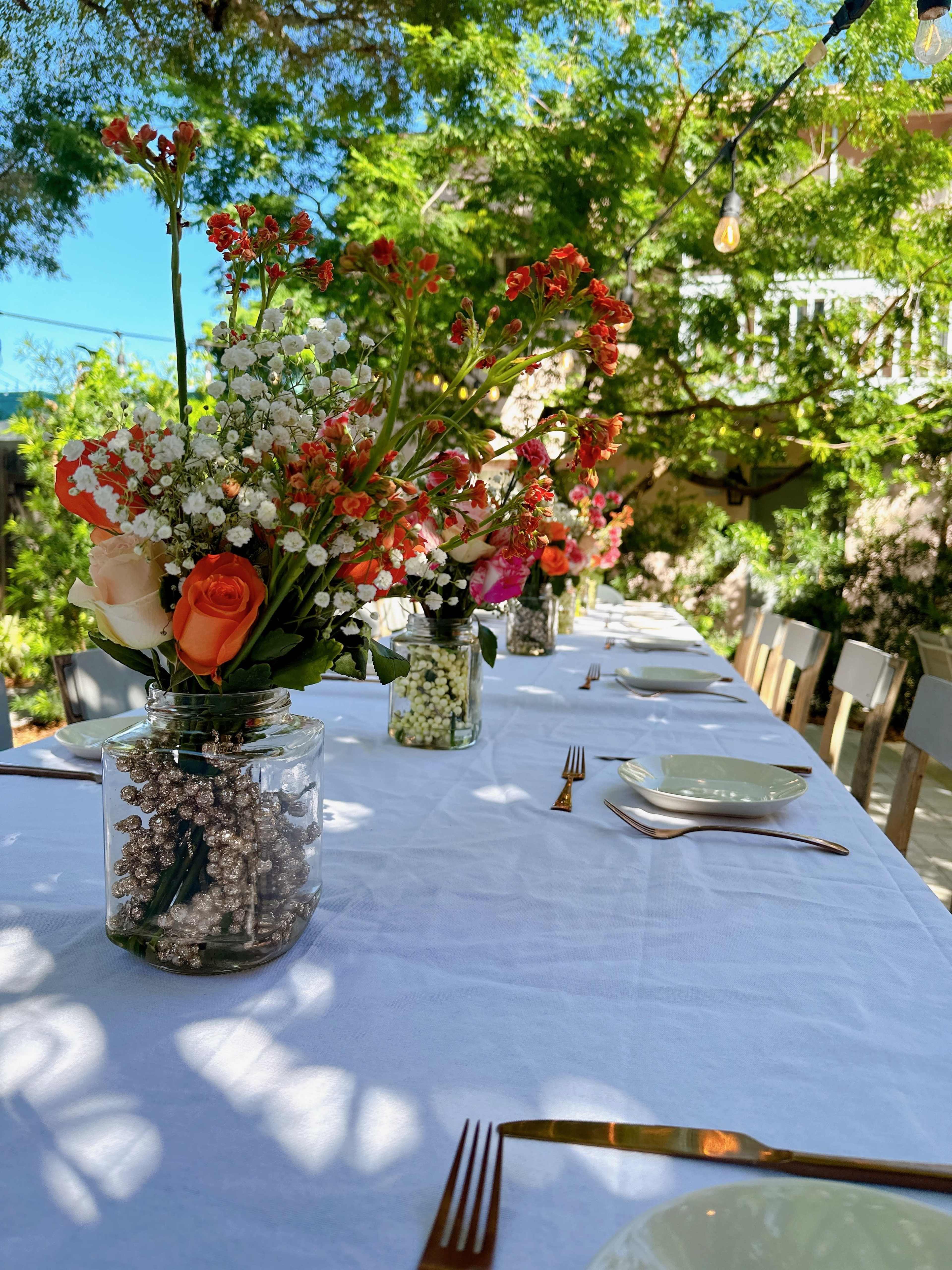 A long outdoor table is set with white tablecloths, adorned with floral centerpieces in glass vases.