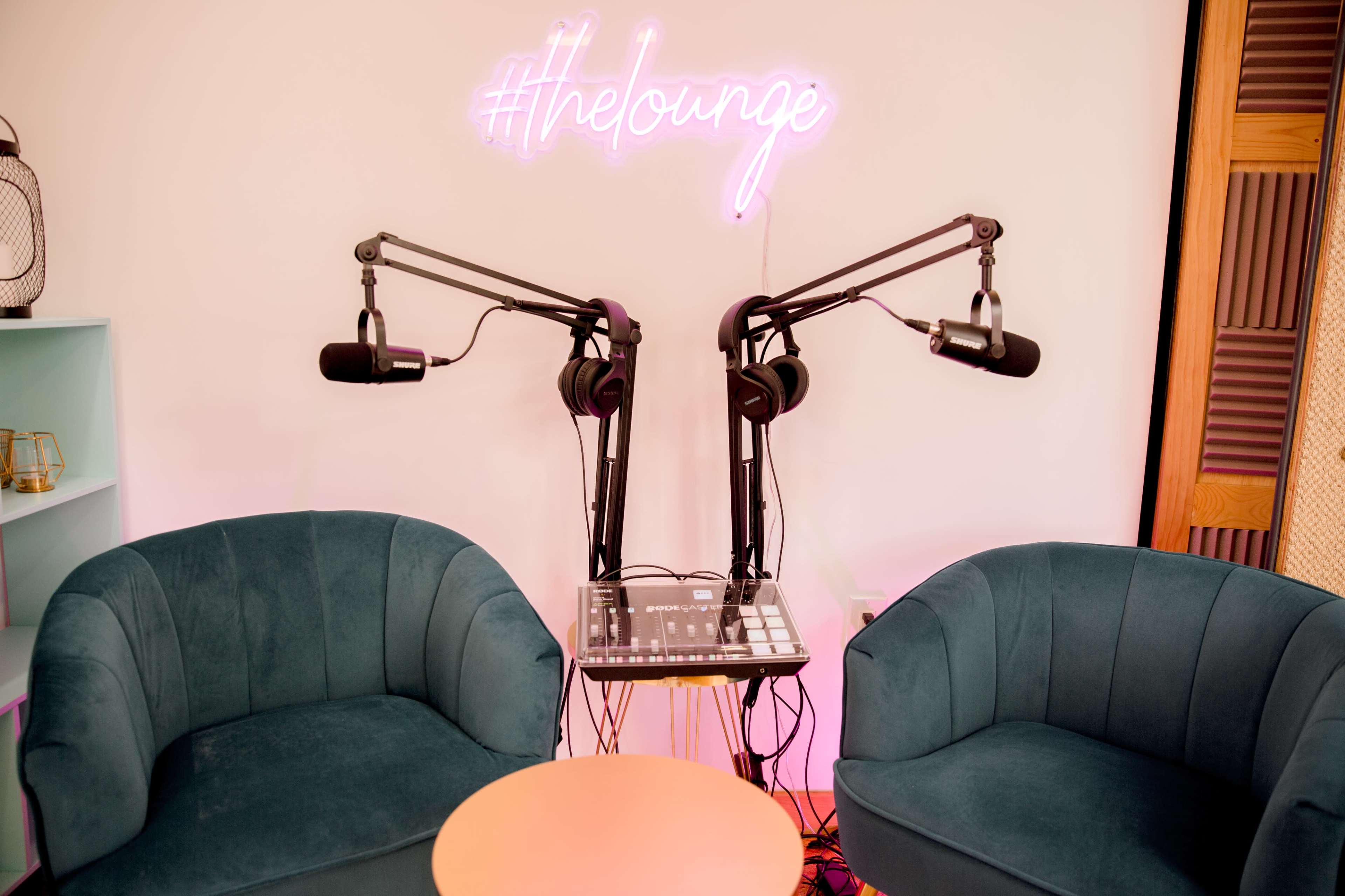 The image shows a podcasting setup with two microphones mounted on adjustable arms, a small table, and a neon sign that says "#thelounge" above a pair of teal chairs.