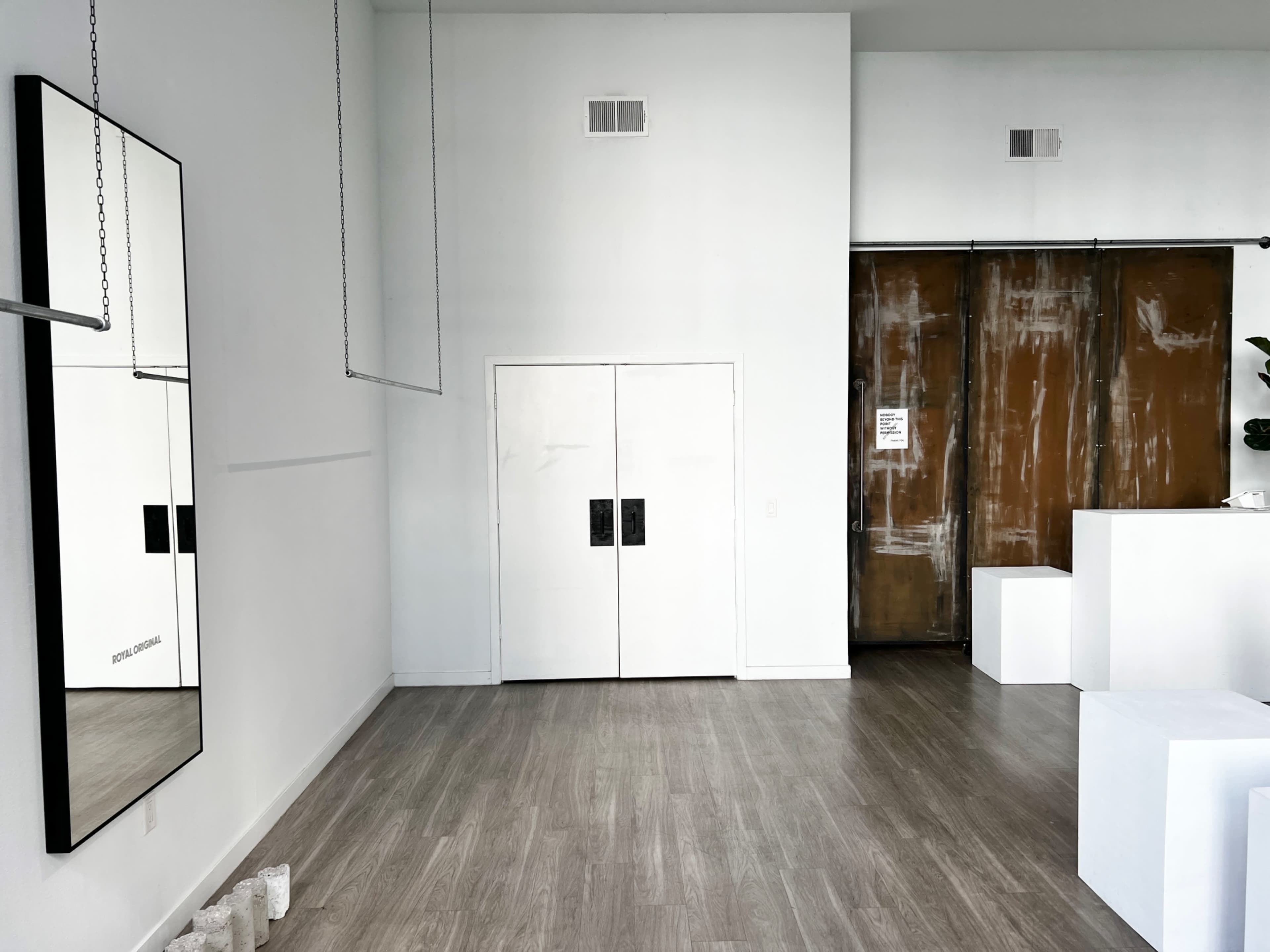 A minimalist interior space with light wood flooring, a large mirror on the left wall, closed double white doors in the center, and a wood-sliding door to the right.