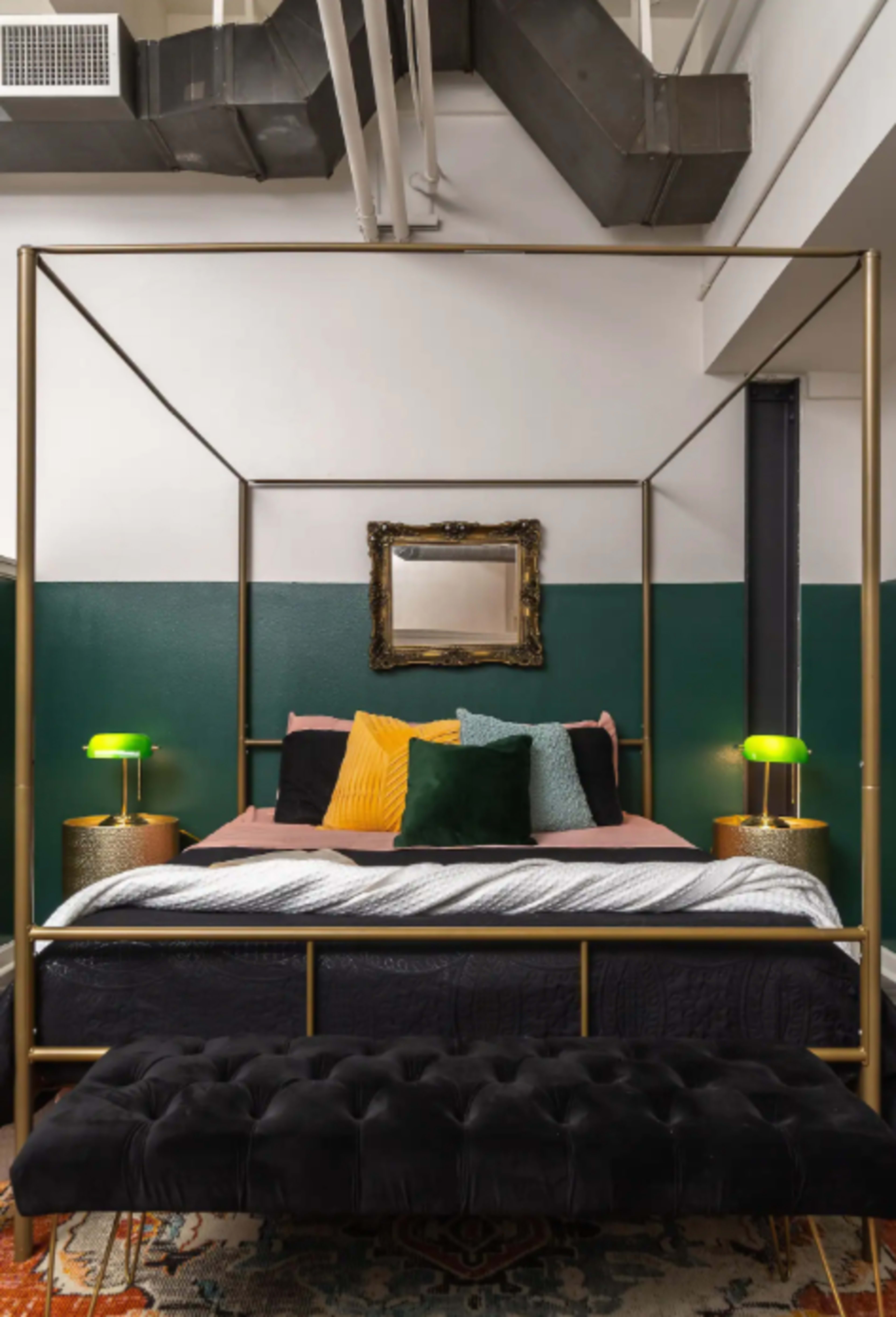 A modern bedroom featuring a four-poster bed with a mix of colorful pillows, two green bedside lamps, and a tufted black bench at the foot of the bed, set against a backdrop of green and white walls.