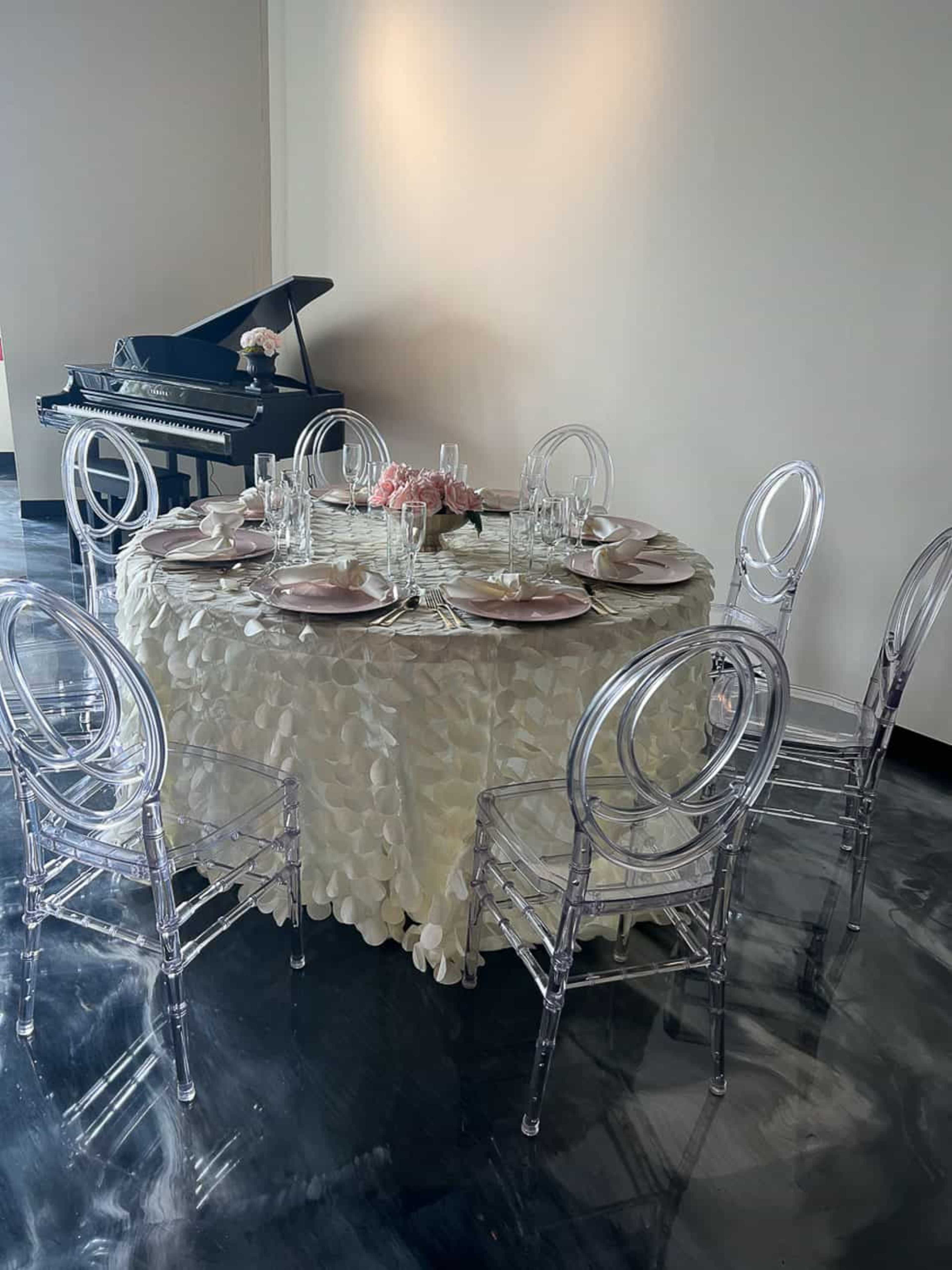 A dining table with a floral tablecloth is set with clear acrylic chairs and surrounded by a grand piano in a well-lit room.