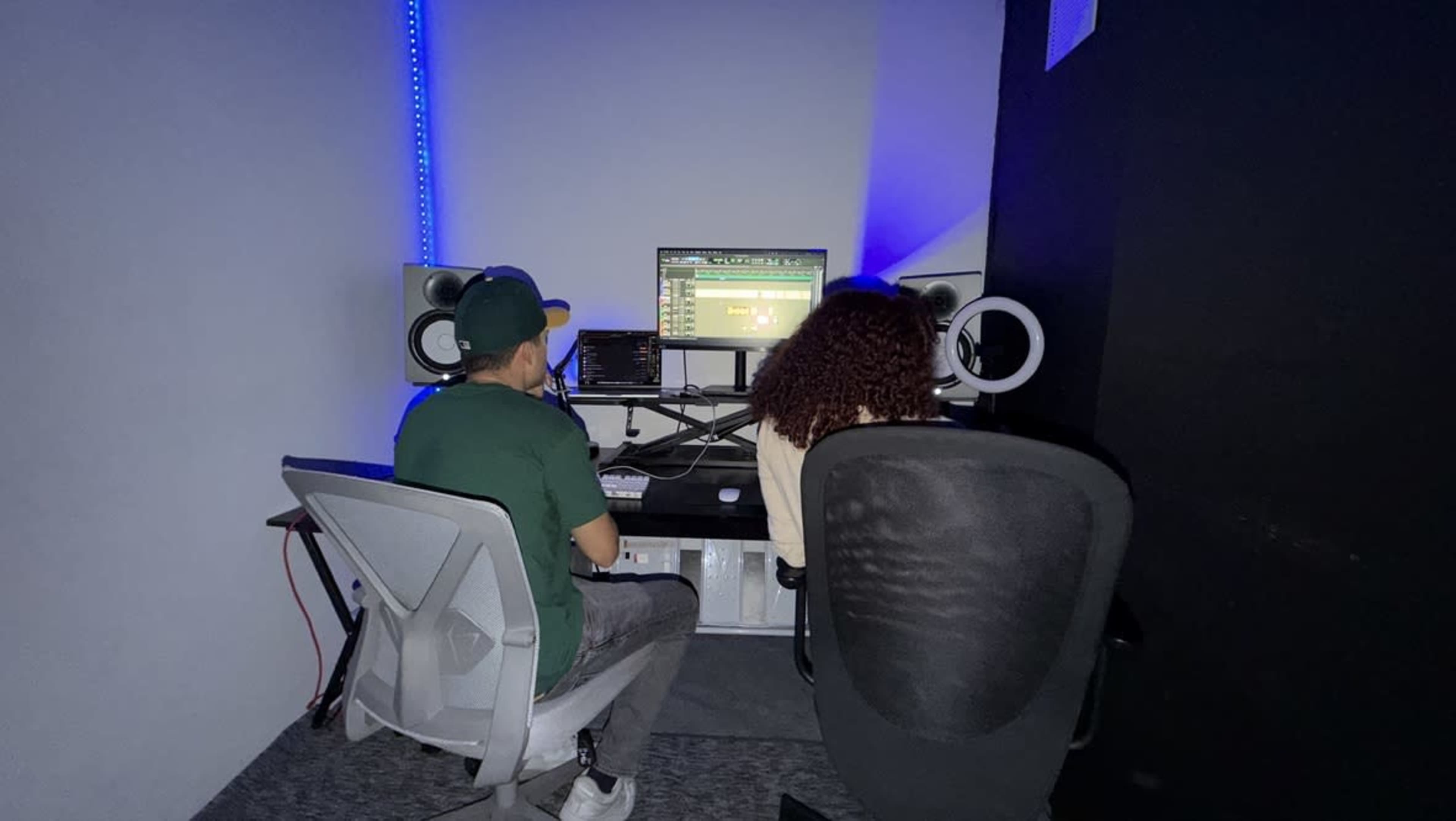 Two individuals are seated in a recording studio, facing a computer with audio software on the screen, surrounded by studio monitors and acoustic treatments.