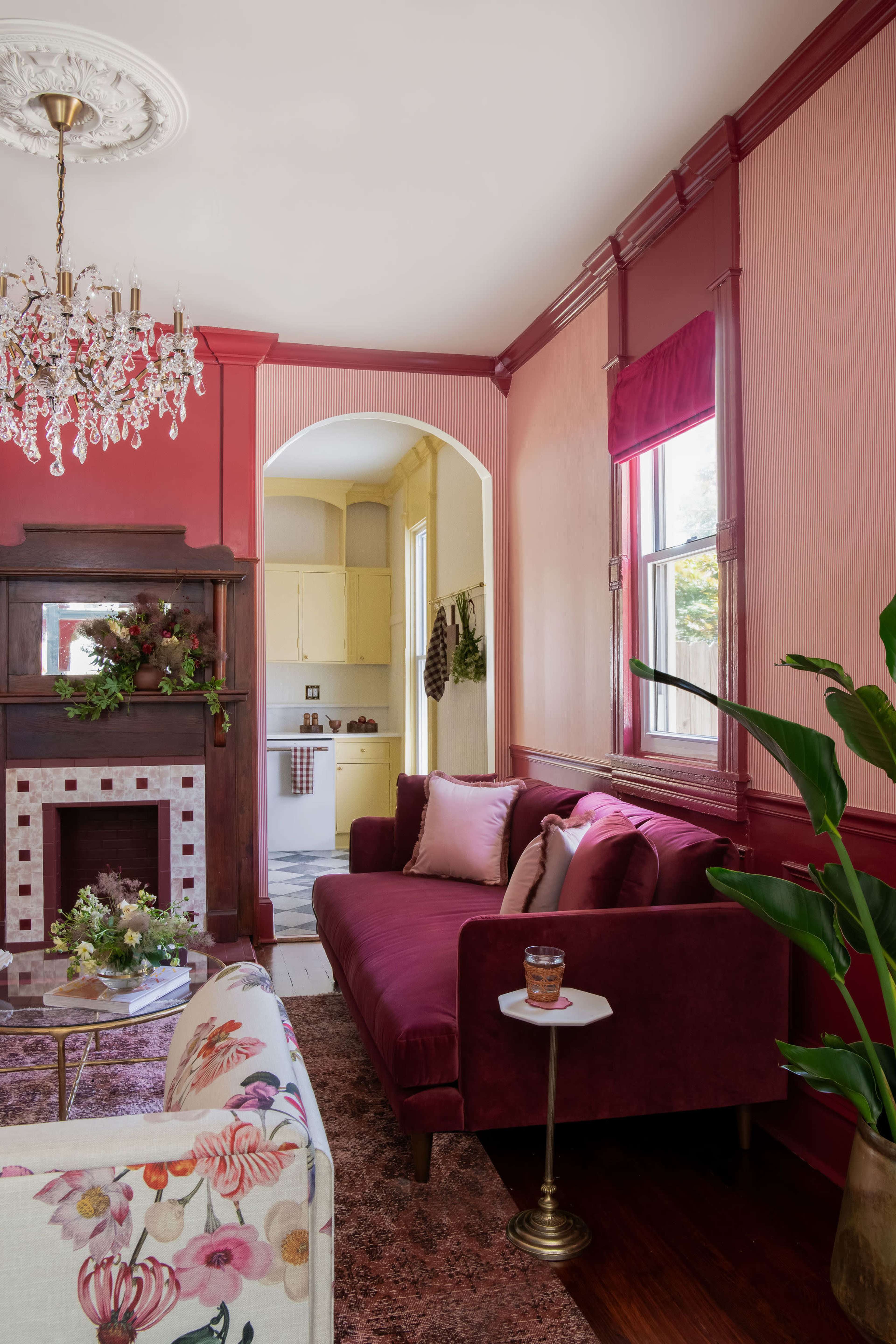 The image shows a cozy living room with a burgundy couch, a floral armchair, and a decorative fireplace, leading to a yellow kitchen visible in the background.