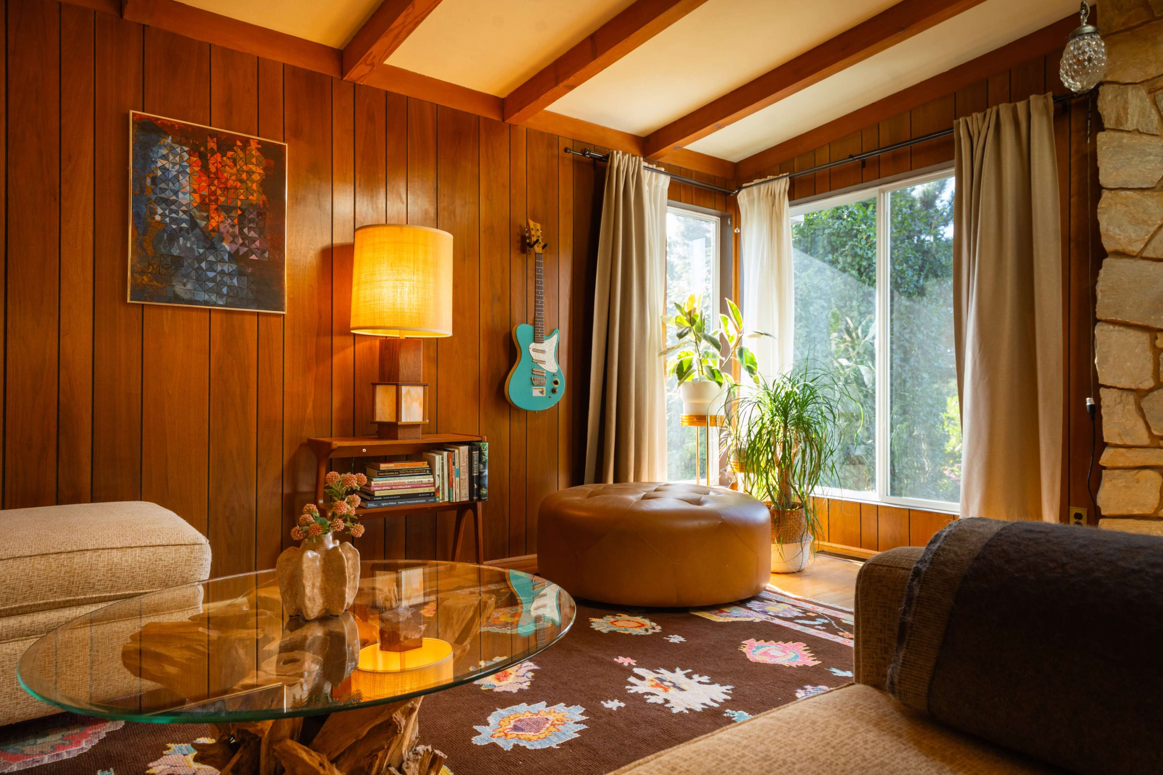The image shows a cozy living room with wooden walls, a large window, an electric guitar on the wall, and a glass coffee table.