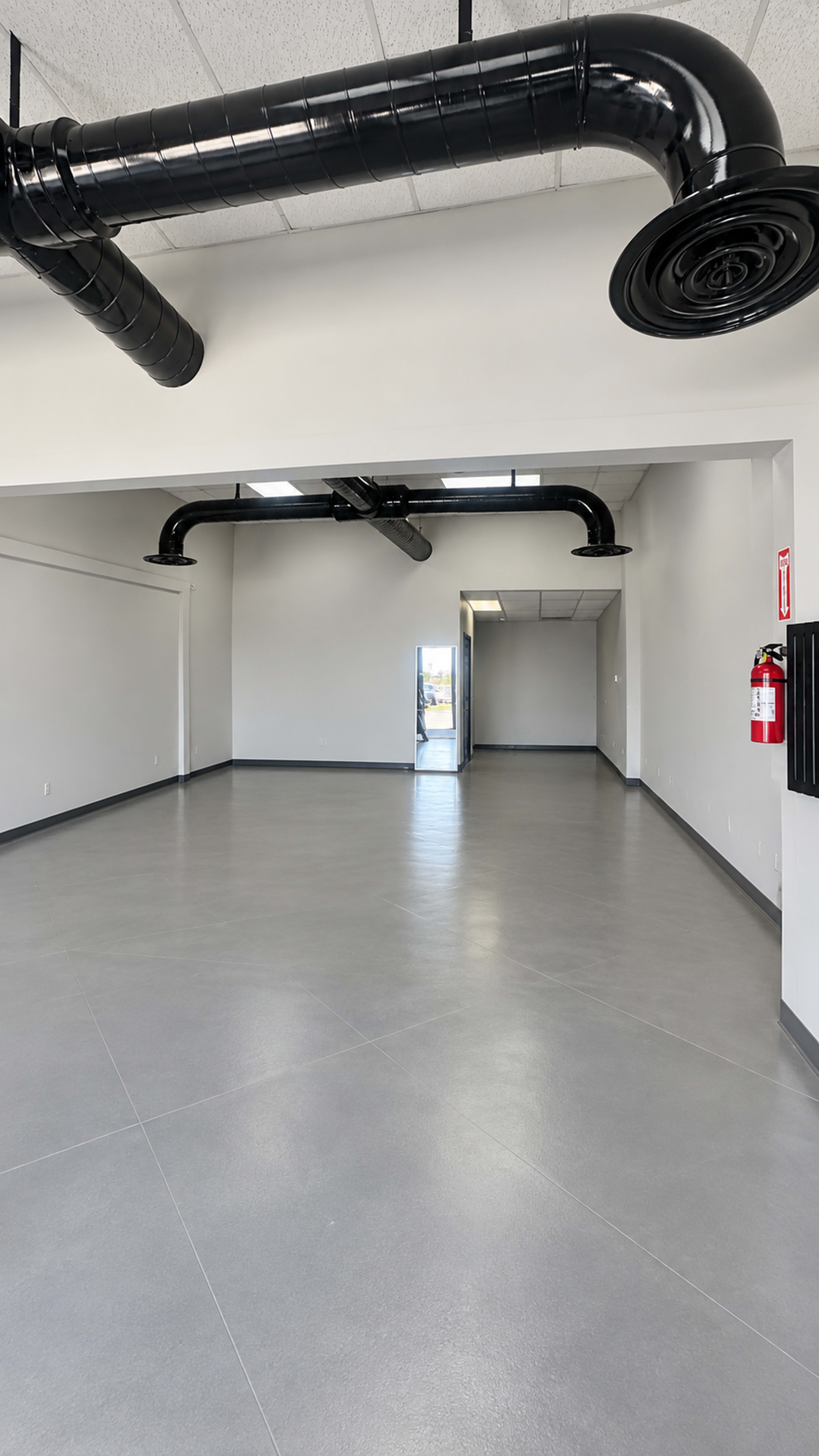 The image shows an empty commercial space with gray flooring and black ductwork along the ceiling.
