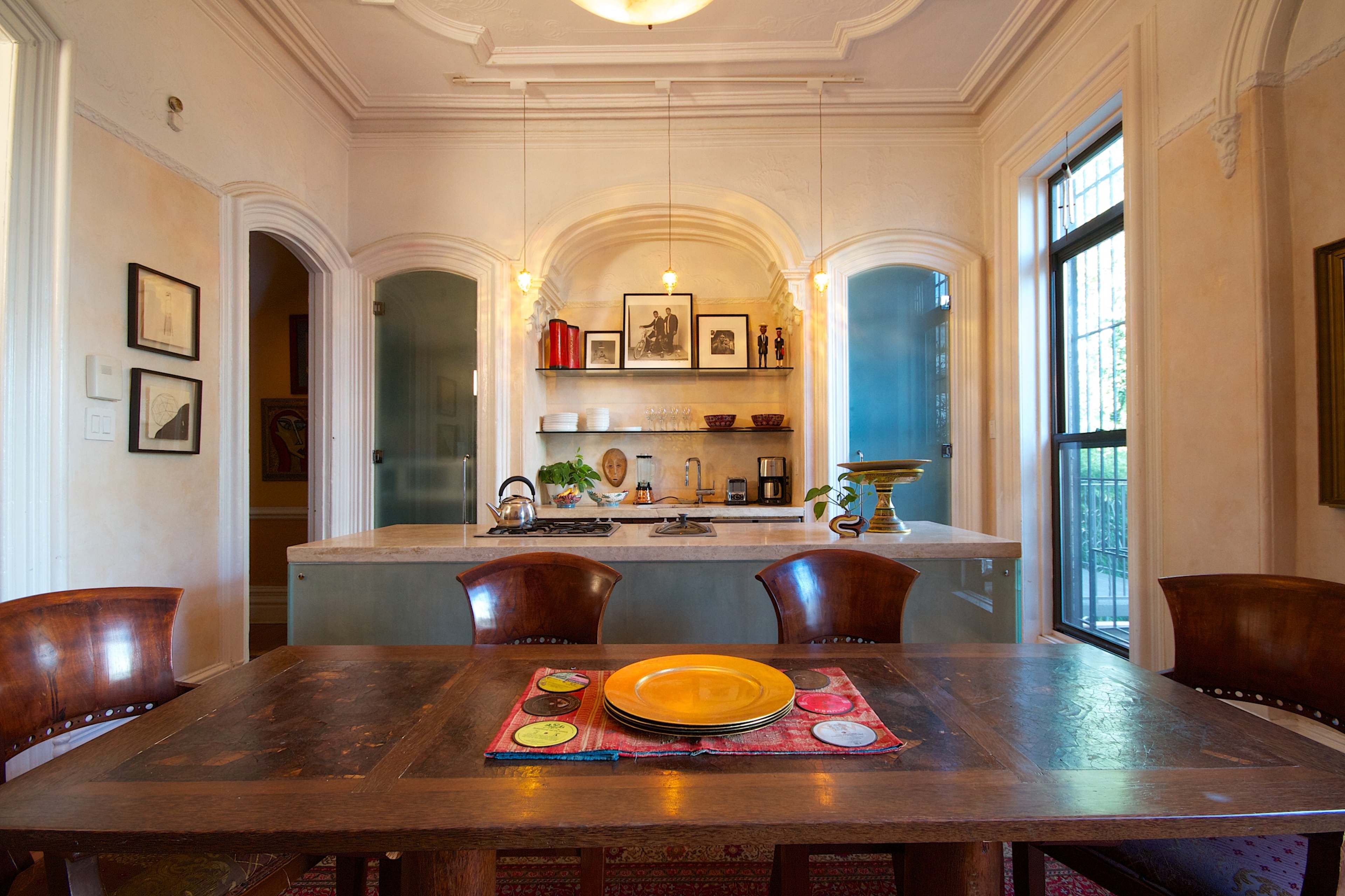 A dining area with a wooden table set with a yellow plate, surrounded by four chairs, and a kitchen area visible in the background.