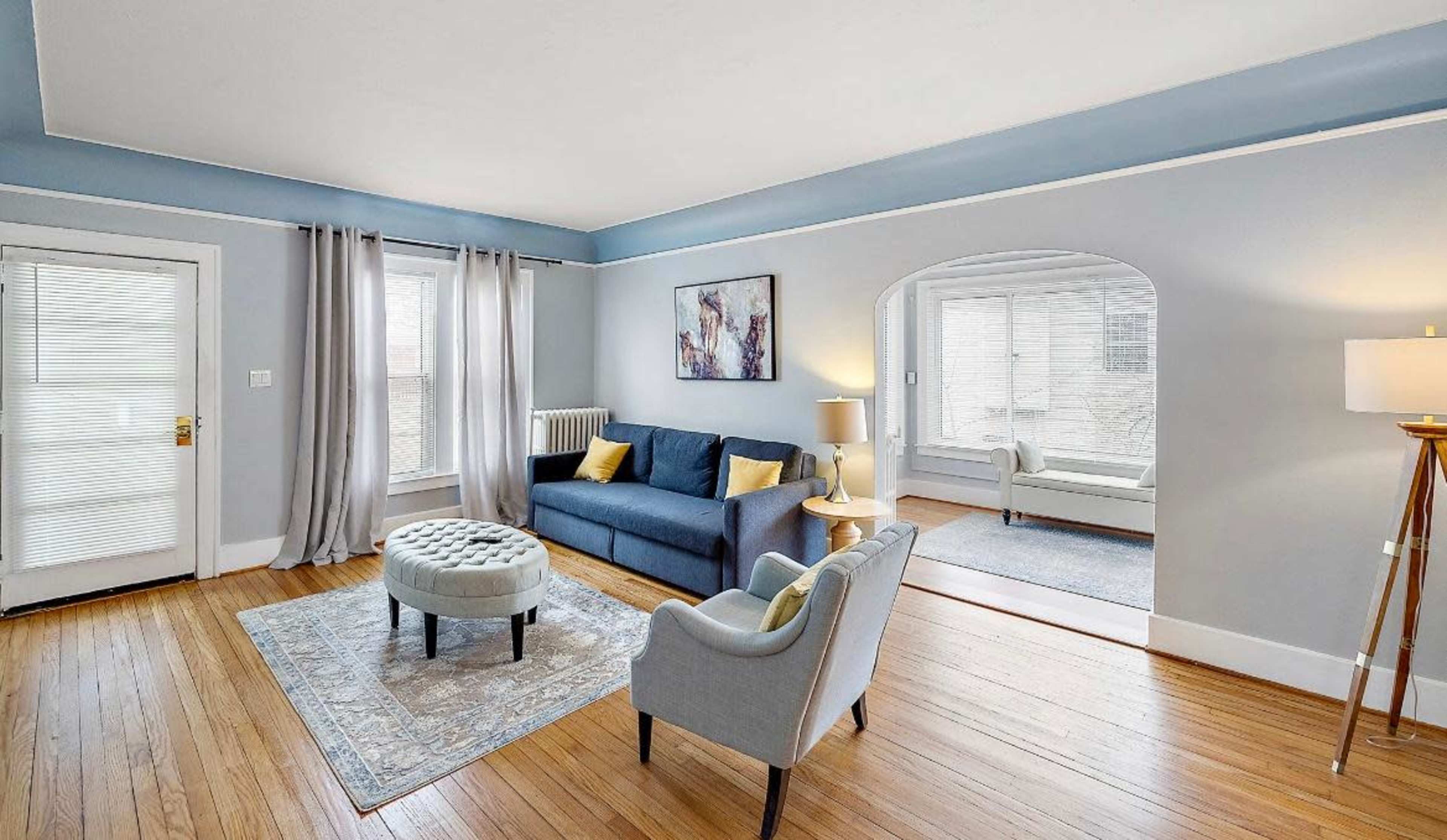 A living room with light blue walls features a sofa, an armchair, a decorative ottoman, and large windows, creating a bright and open atmosphere.