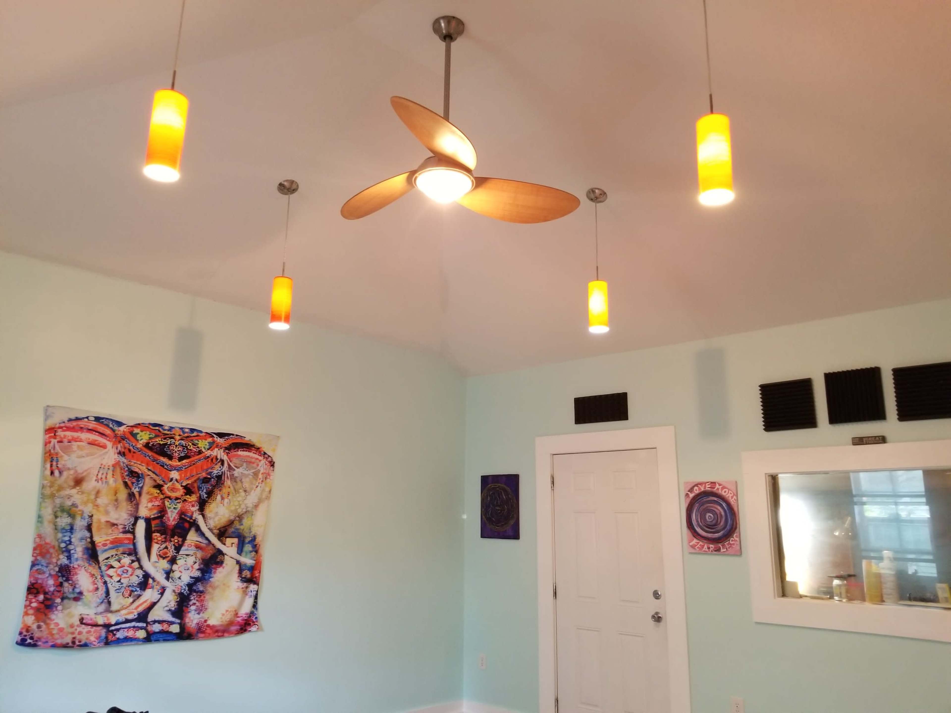 A room with a light-colored wall, a ceiling fan with wooden blades, four hanging light fixtures, and various wall decorations, including a tapestry of an elephant.