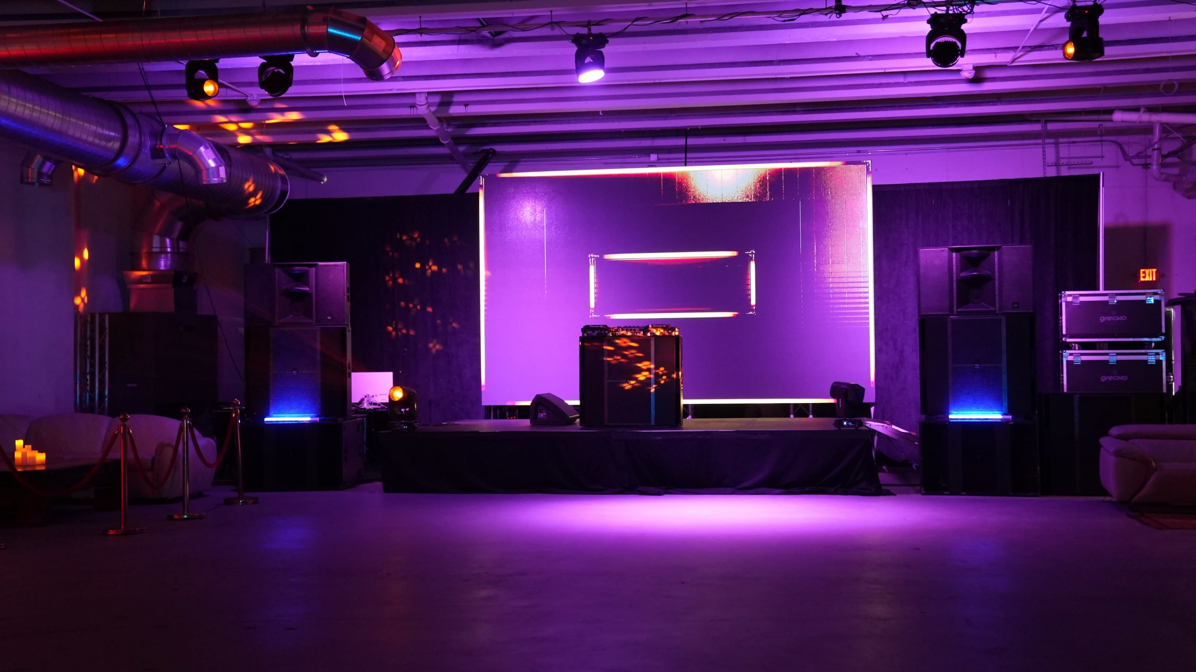 The image shows an empty event space with a stage featuring speakers and a large screen, illuminated by purple and orange lighting.