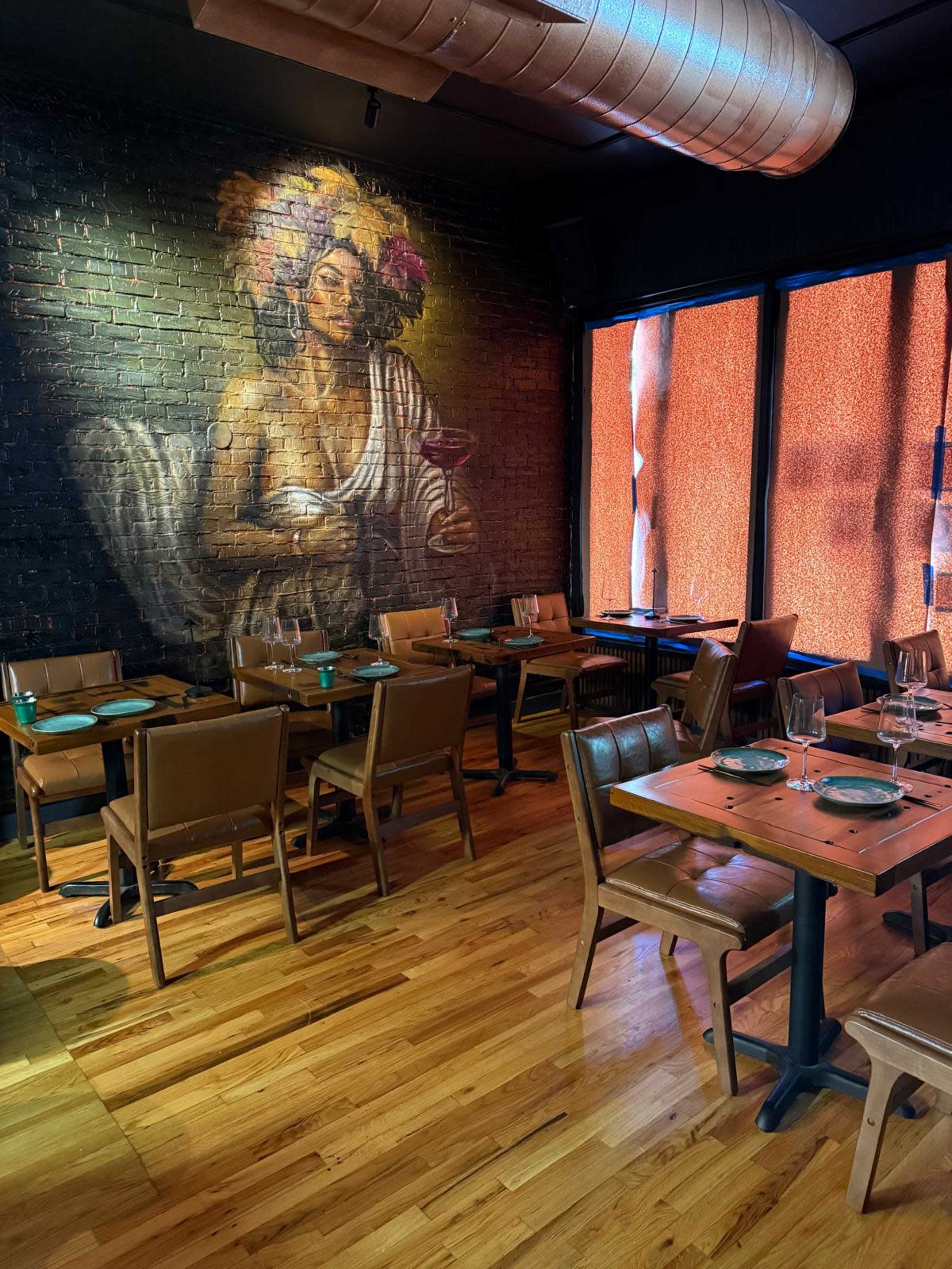 The image shows a dining area with wooden tables and chairs, featuring a large mural of a woman on the brick wall and orange-tinted windows.