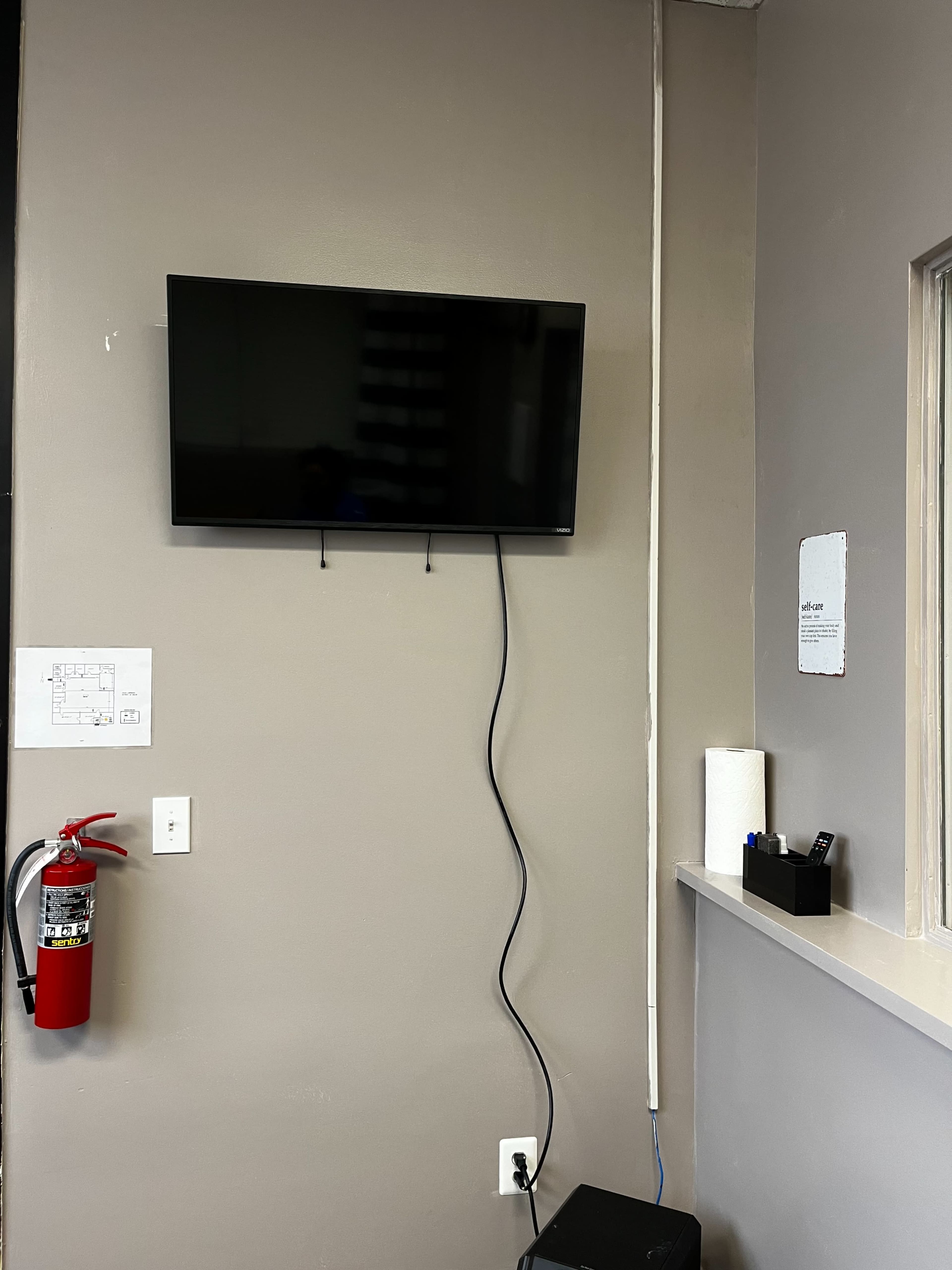 A flat-screen television is mounted on a gray wall, with a fire extinguisher and a small black box on a counter below.