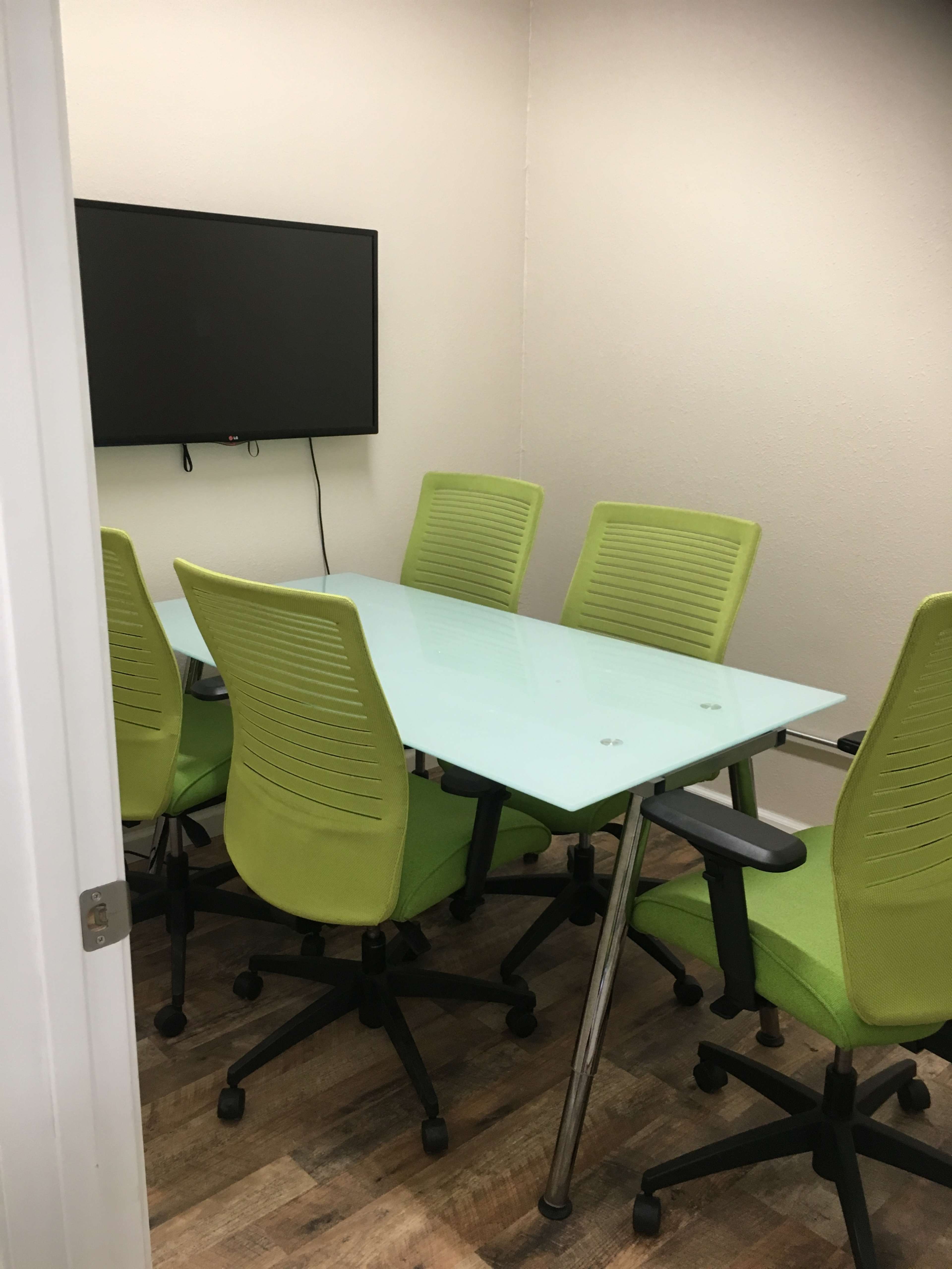 A glass conference table surrounded by six green chairs sits in a small, well-lit meeting room with a wall-mounted screen.