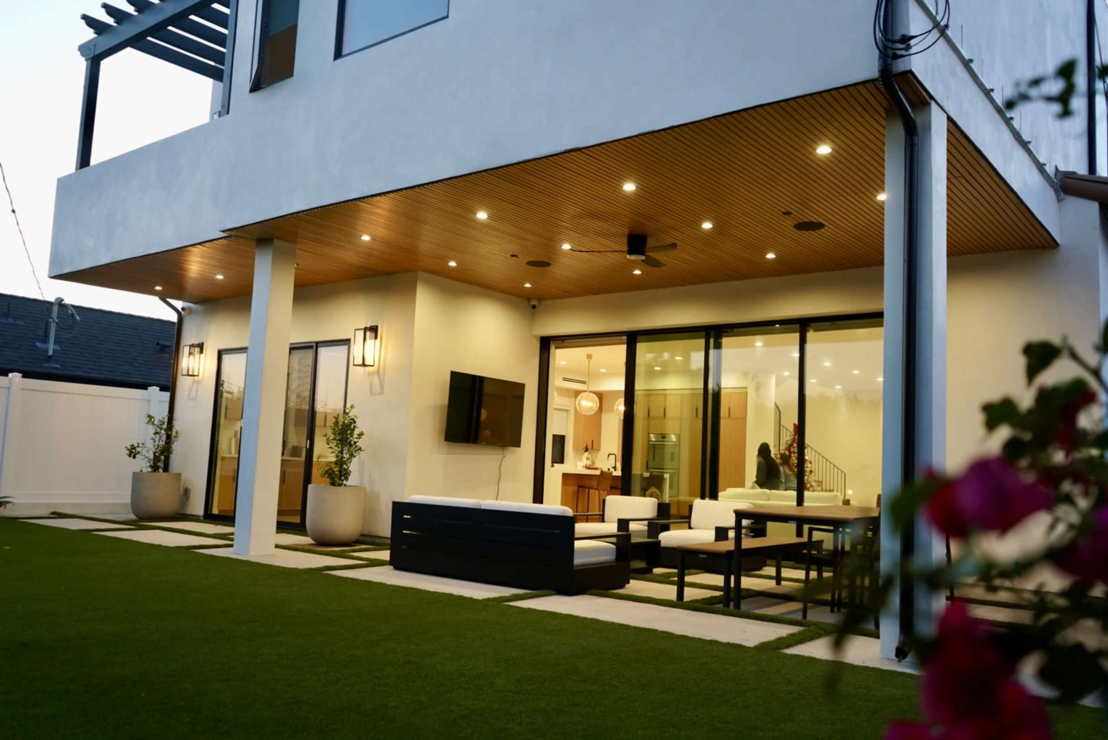 The image shows a modern backyard patio with a seating area, large glass doors leading into a well-lit interior space, and manicured green grass.