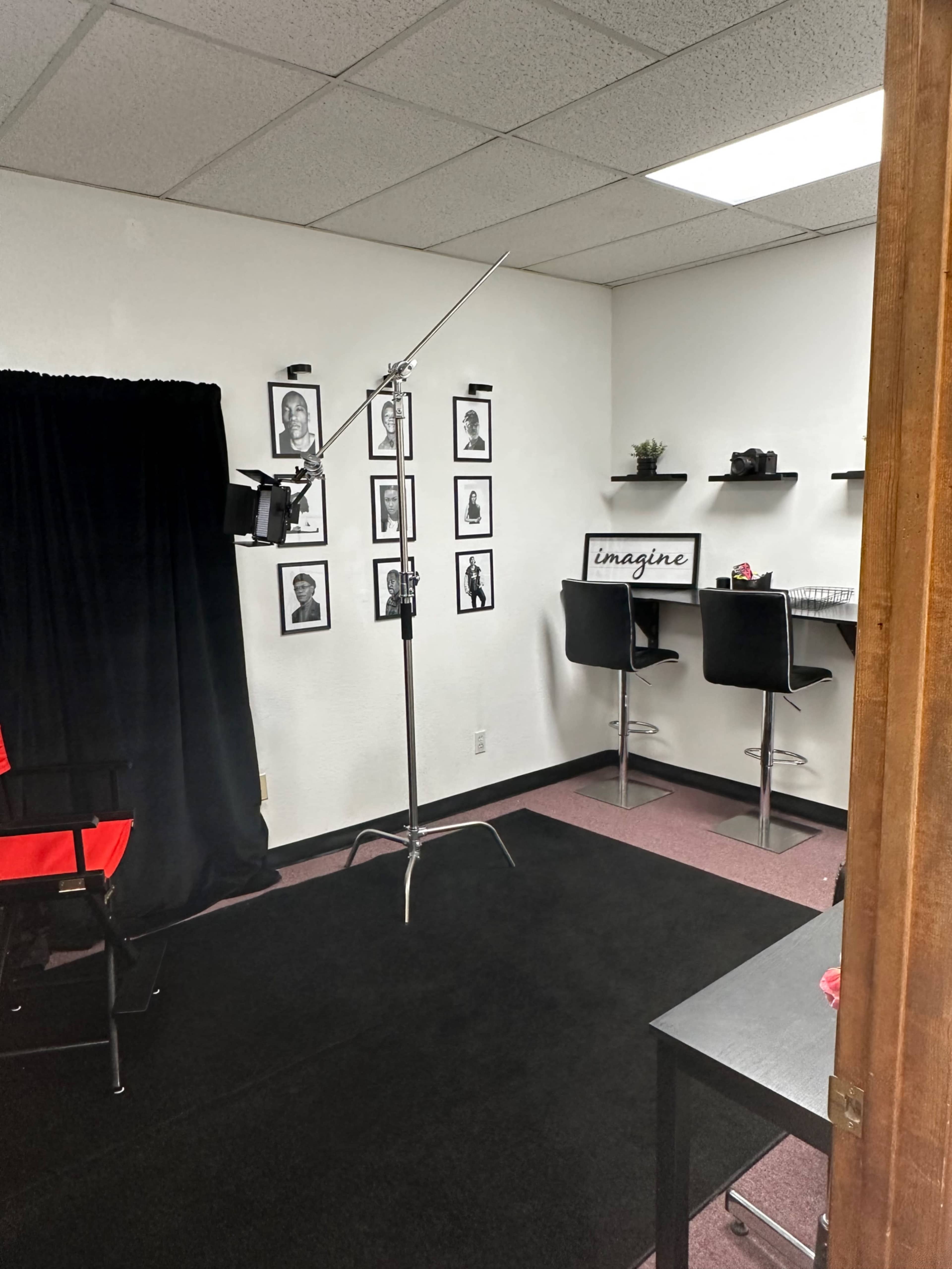 A small studio space with a black backdrop, framed photographs on the wall, a standing light on a stand, and a small counter with chairs.