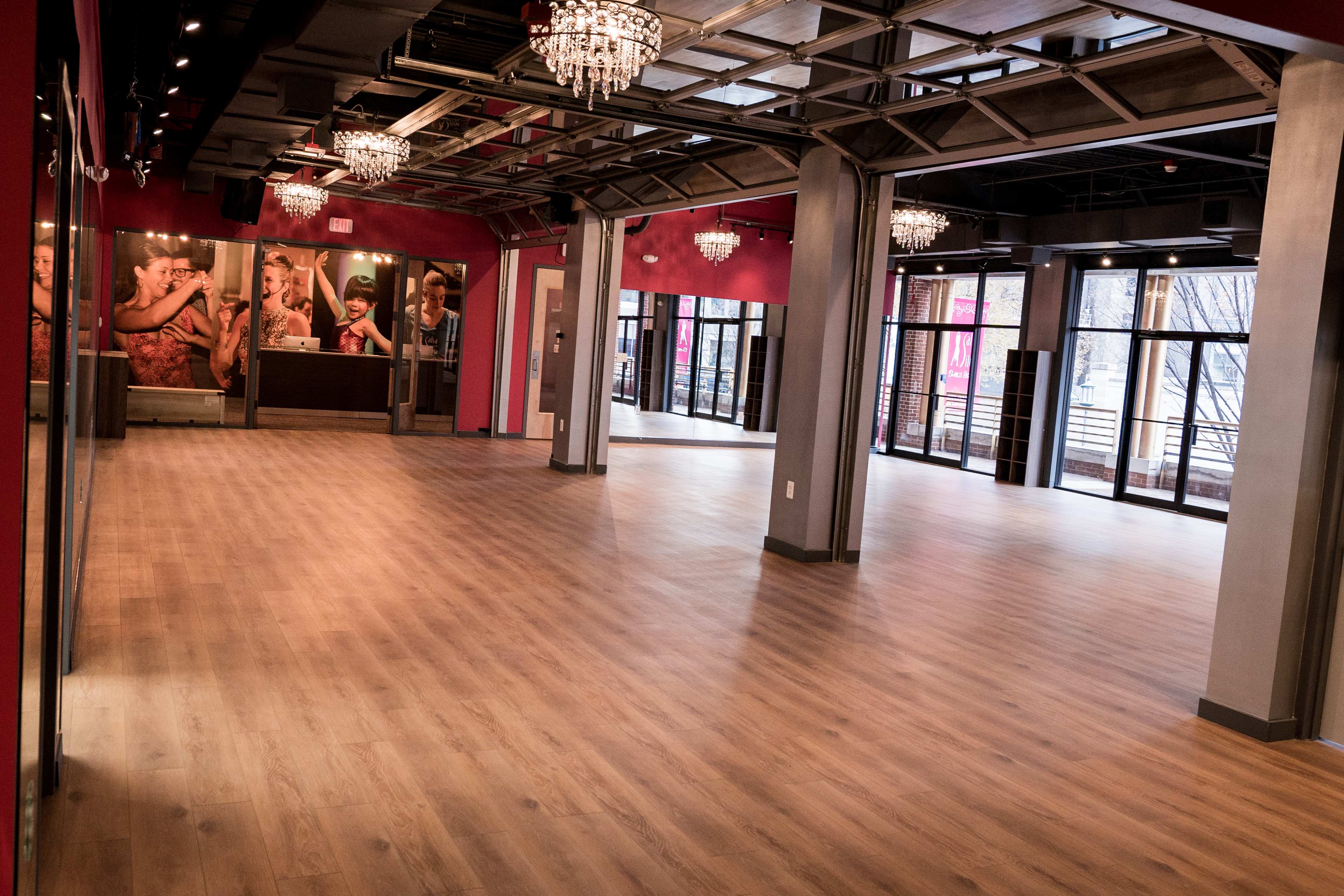 A spacious, empty room with hardwood flooring, large windows, and chandeliers, accented by wall art featuring people.