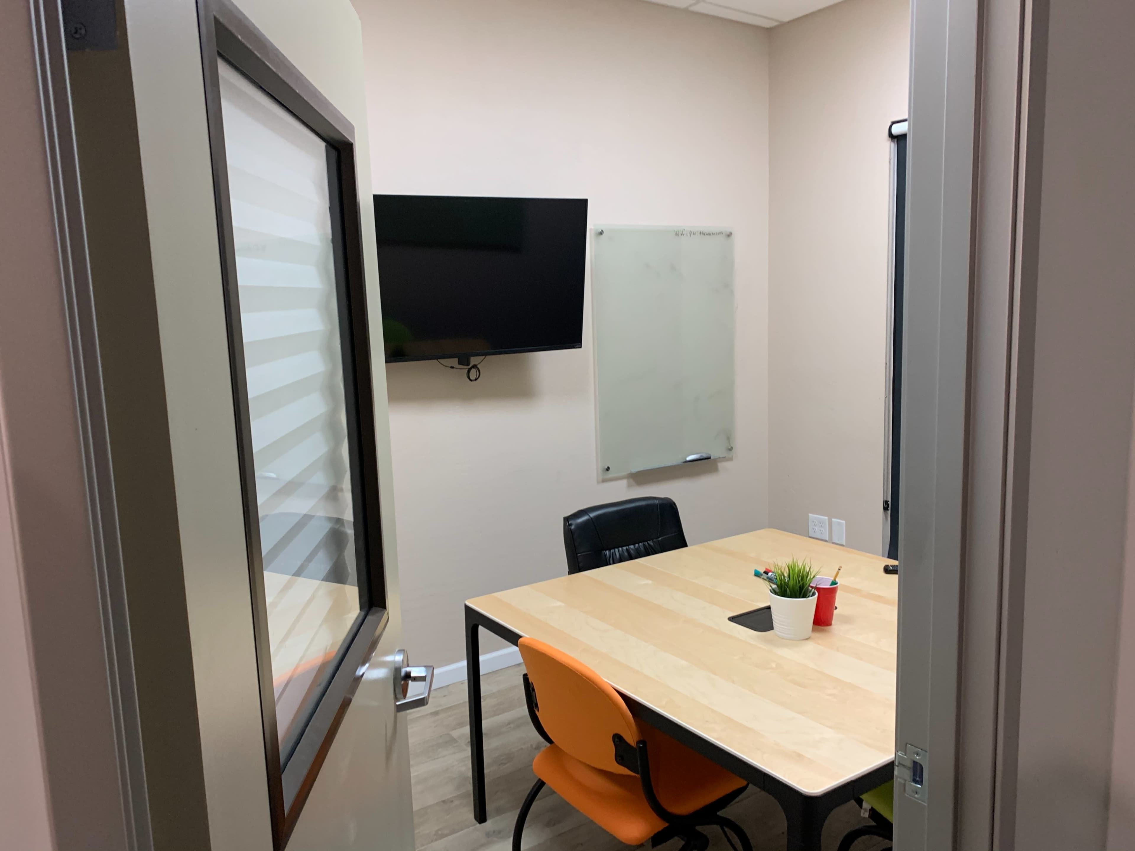 A small conference room with a wooden table, two orange chairs, a black chair, a wall-mounted television, and a whiteboard.