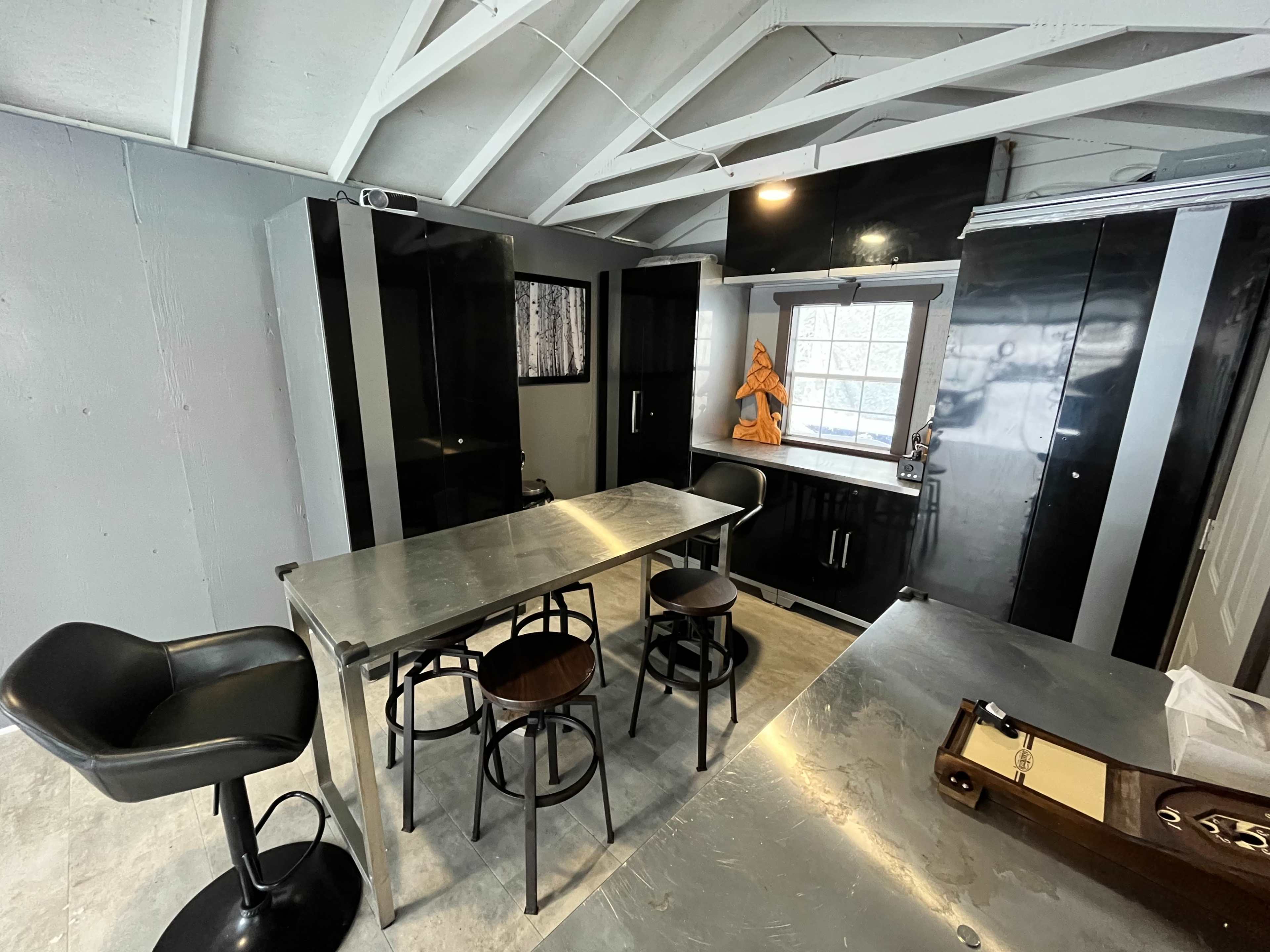 A modern kitchen features sleek black cabinets, a metallic table, and bar stools under a slanted ceiling with exposed beams.