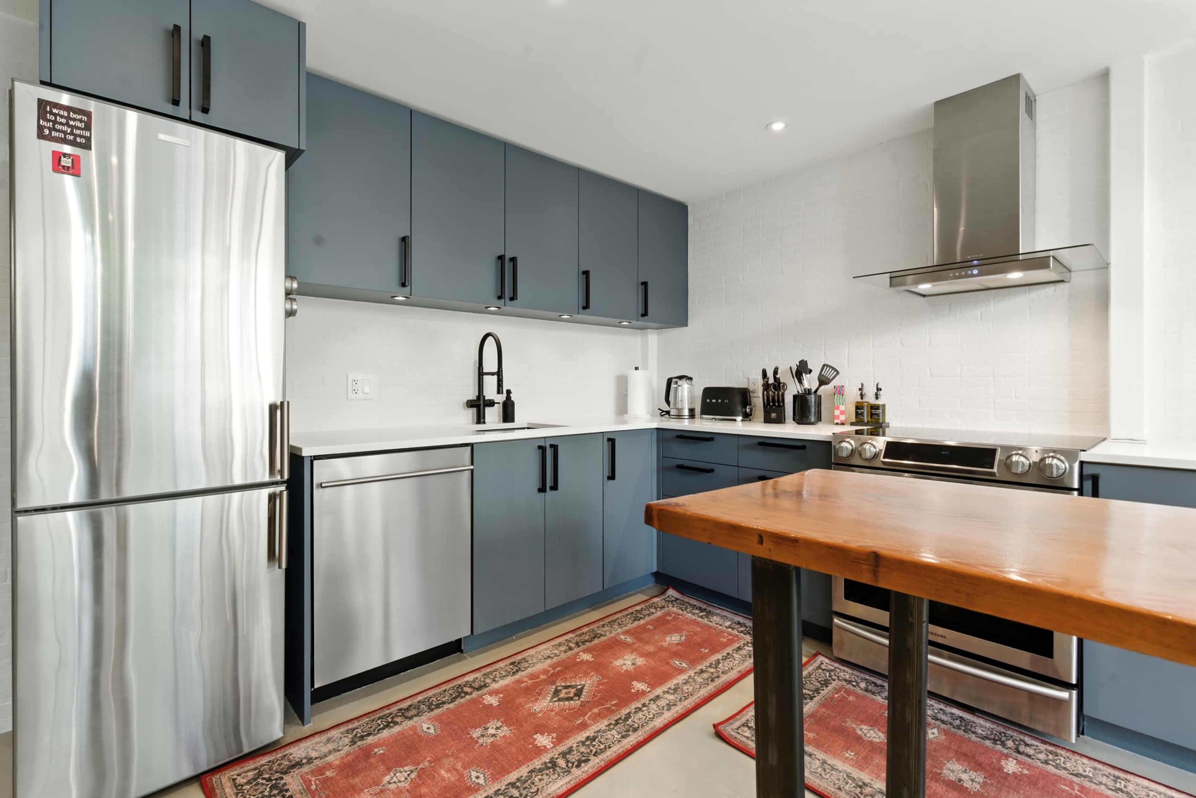 A modern kitchen features blue cabinets, stainless steel appliances, and a wooden island with a red patterned rug beneath it.