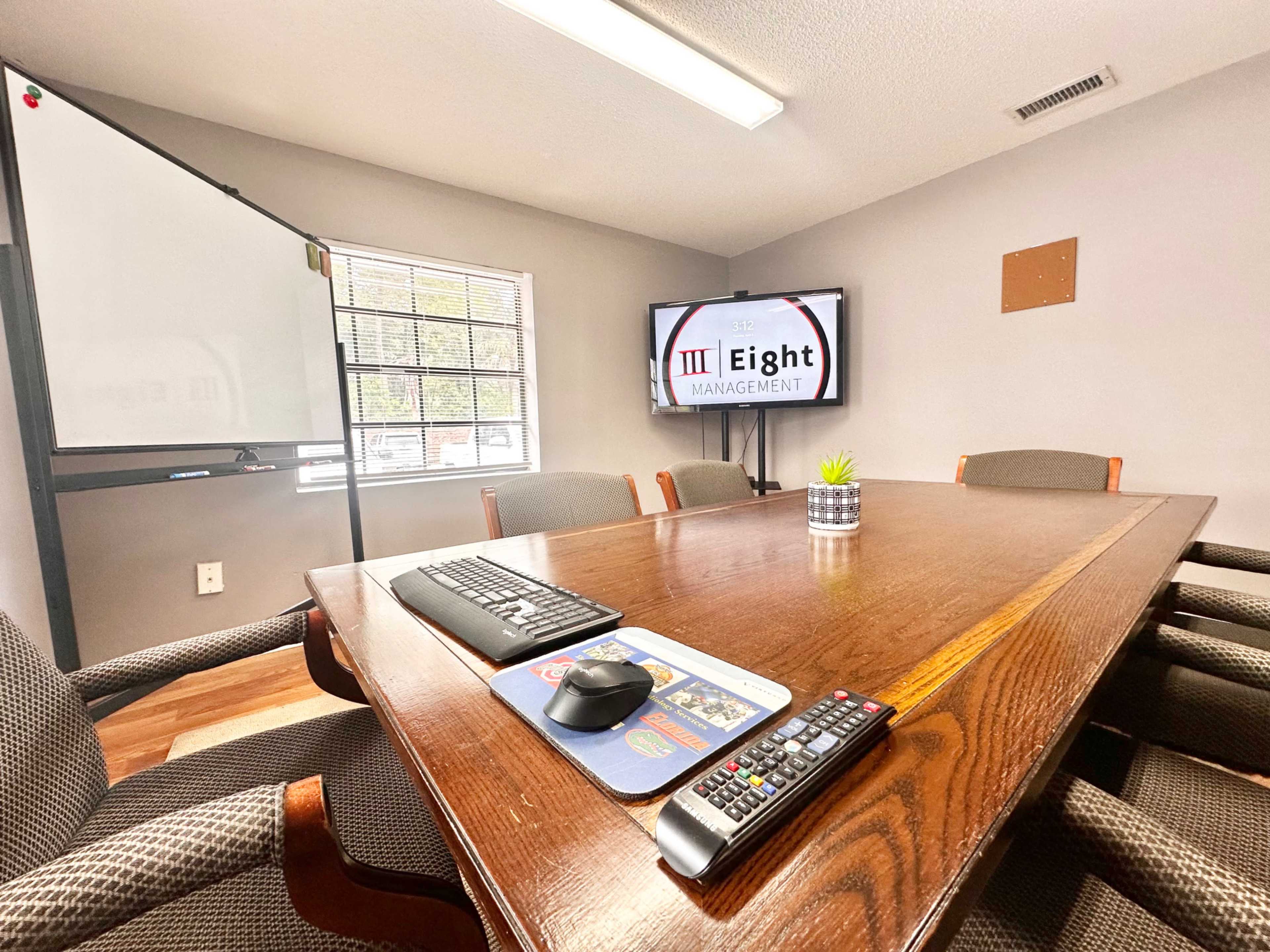 The image shows a conference room featuring a large wooden table, several chairs, a wall-mounted television displaying "Eight Management," and a whiteboard in the background.
