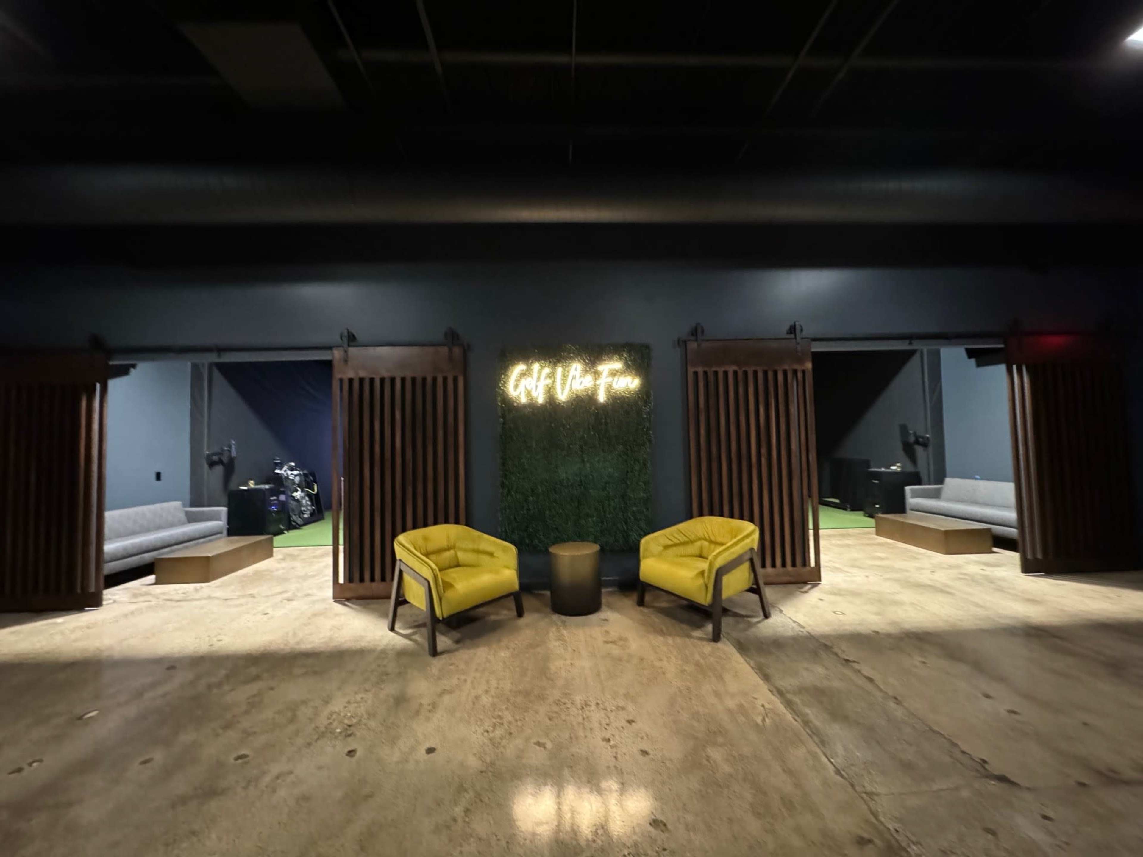 The image shows a modern lounge area with two yellow armchairs facing a green wall featuring the text "Golf & Fun," with wooden partitions leading to darker rooms on either side.