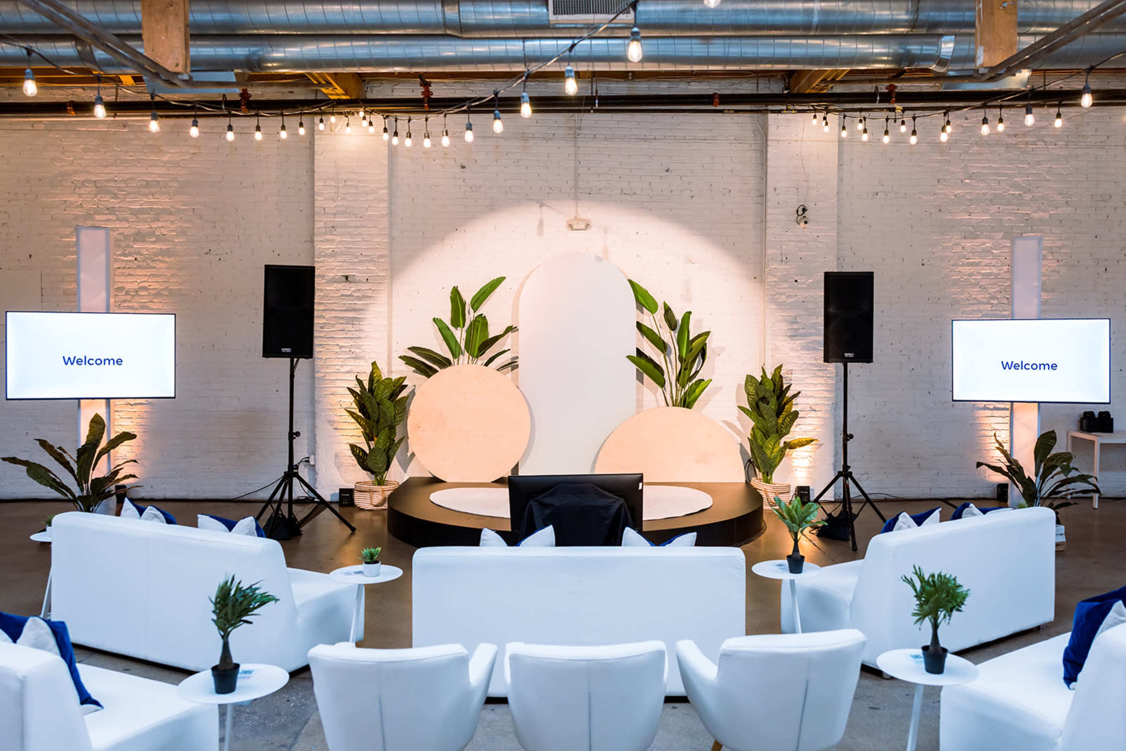 The image shows a modern event venue with white couches arranged in a semicircle facing a circular stage, flanked by large potted plants and screens displaying the word "Welcome."