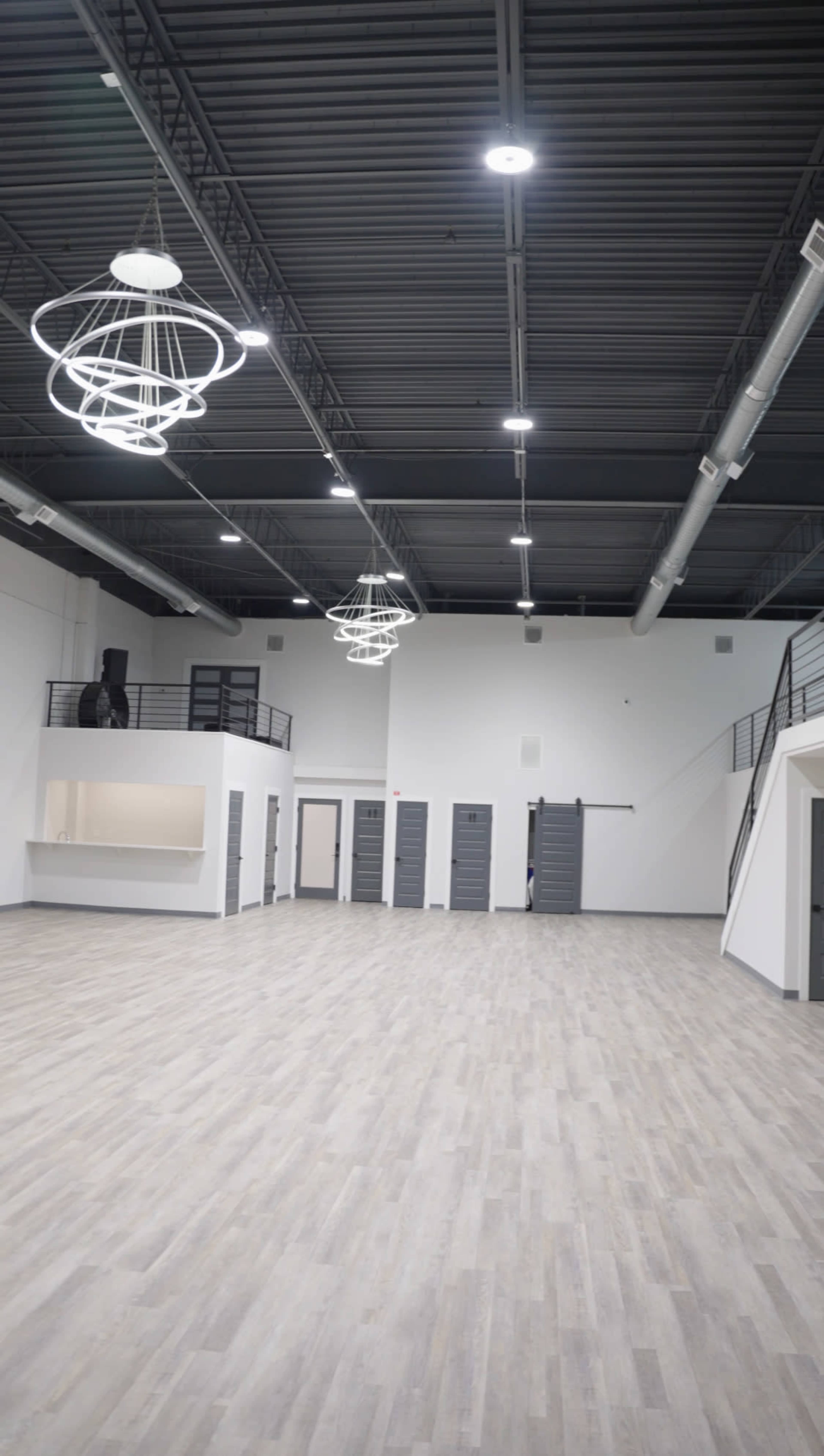 The image shows a spacious, modern interior with wooden flooring, white walls, and multiple industrial-style light fixtures hanging from the ceiling.