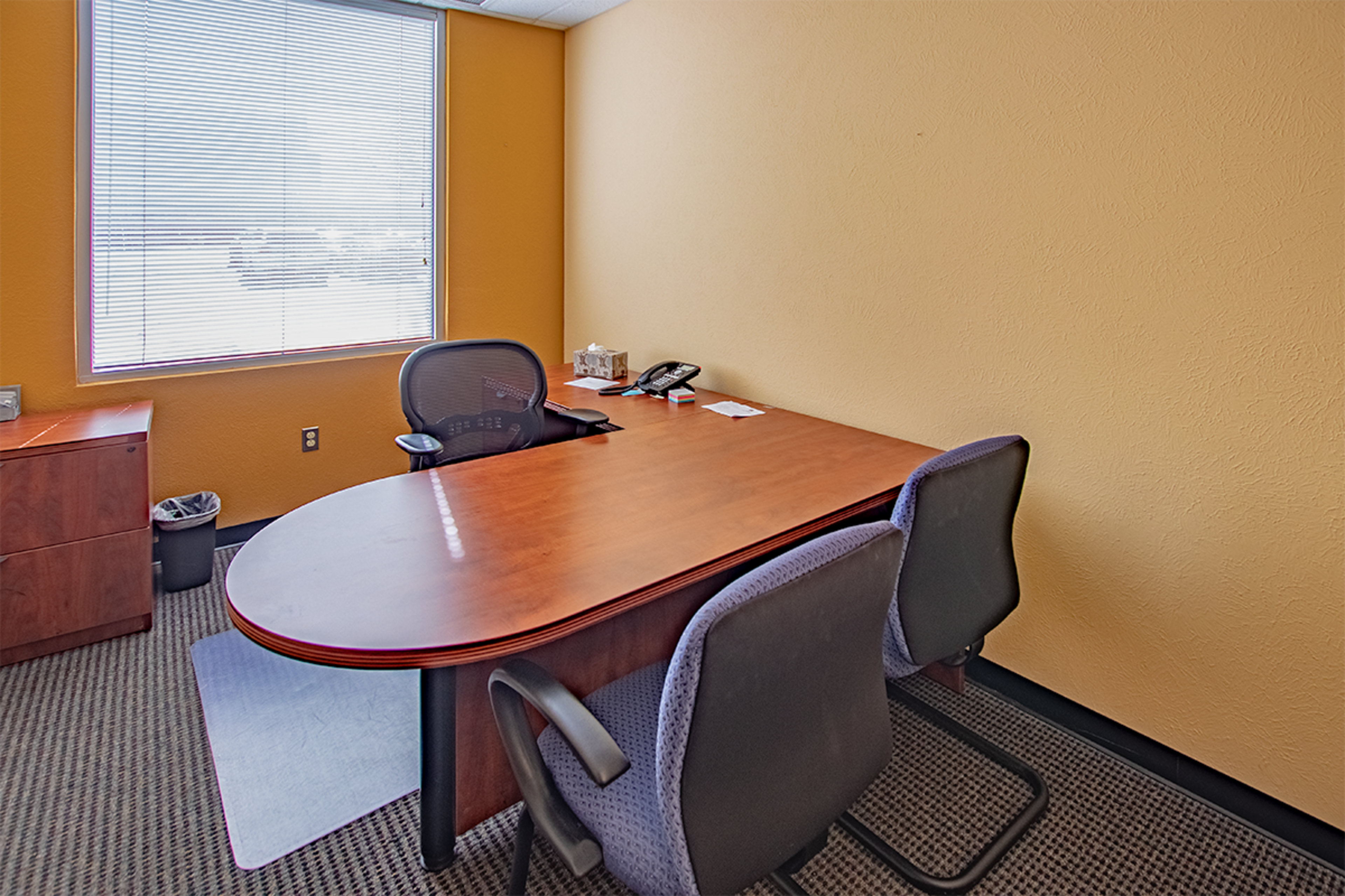 A small office featuring a wooden desk with two chairs, a telephone, and a window with blinds.