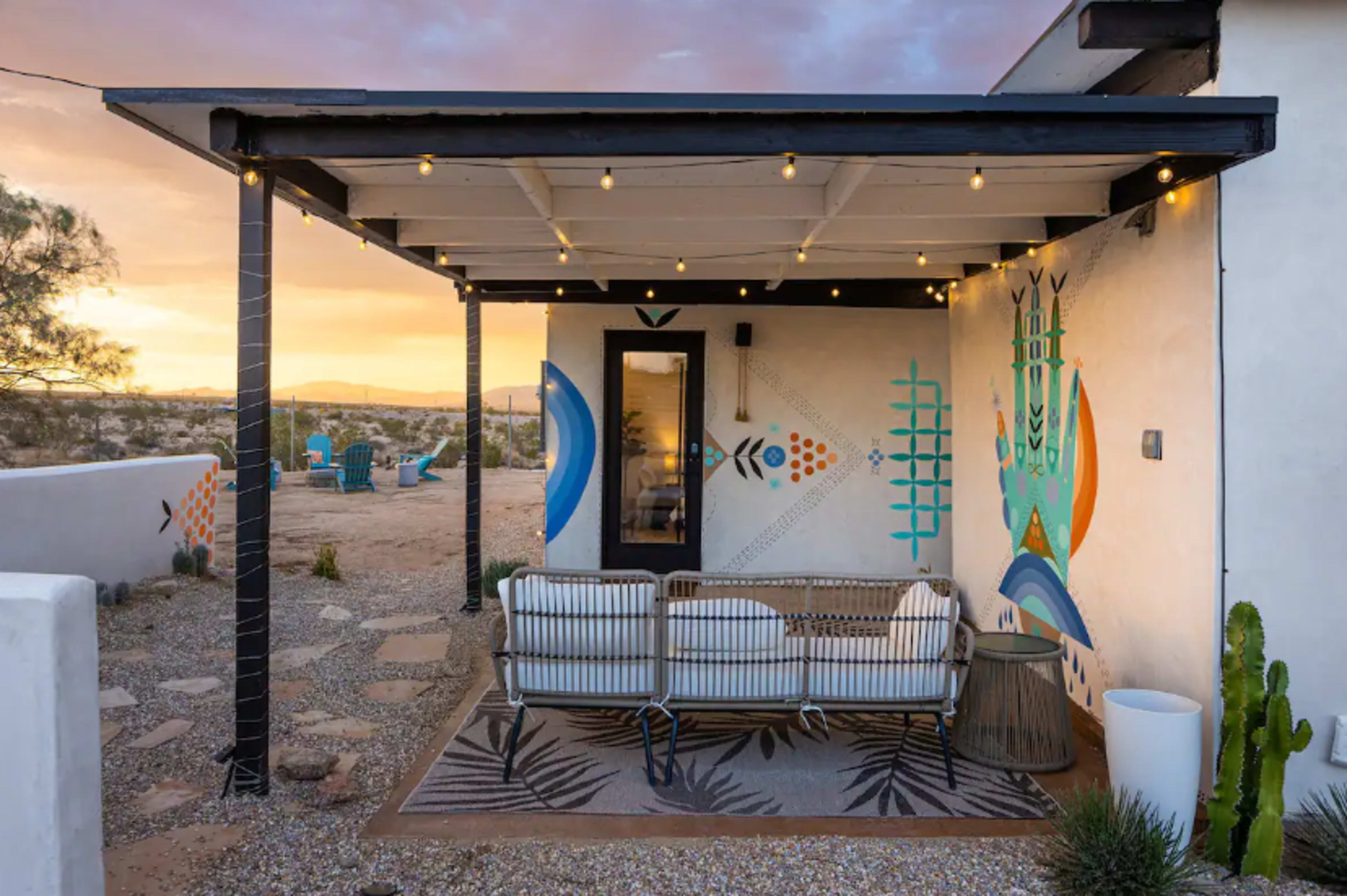 The image shows a patio with a shaded seating area, decorated with colorful murals, and surrounded by a gravel landscape under a sunset sky.