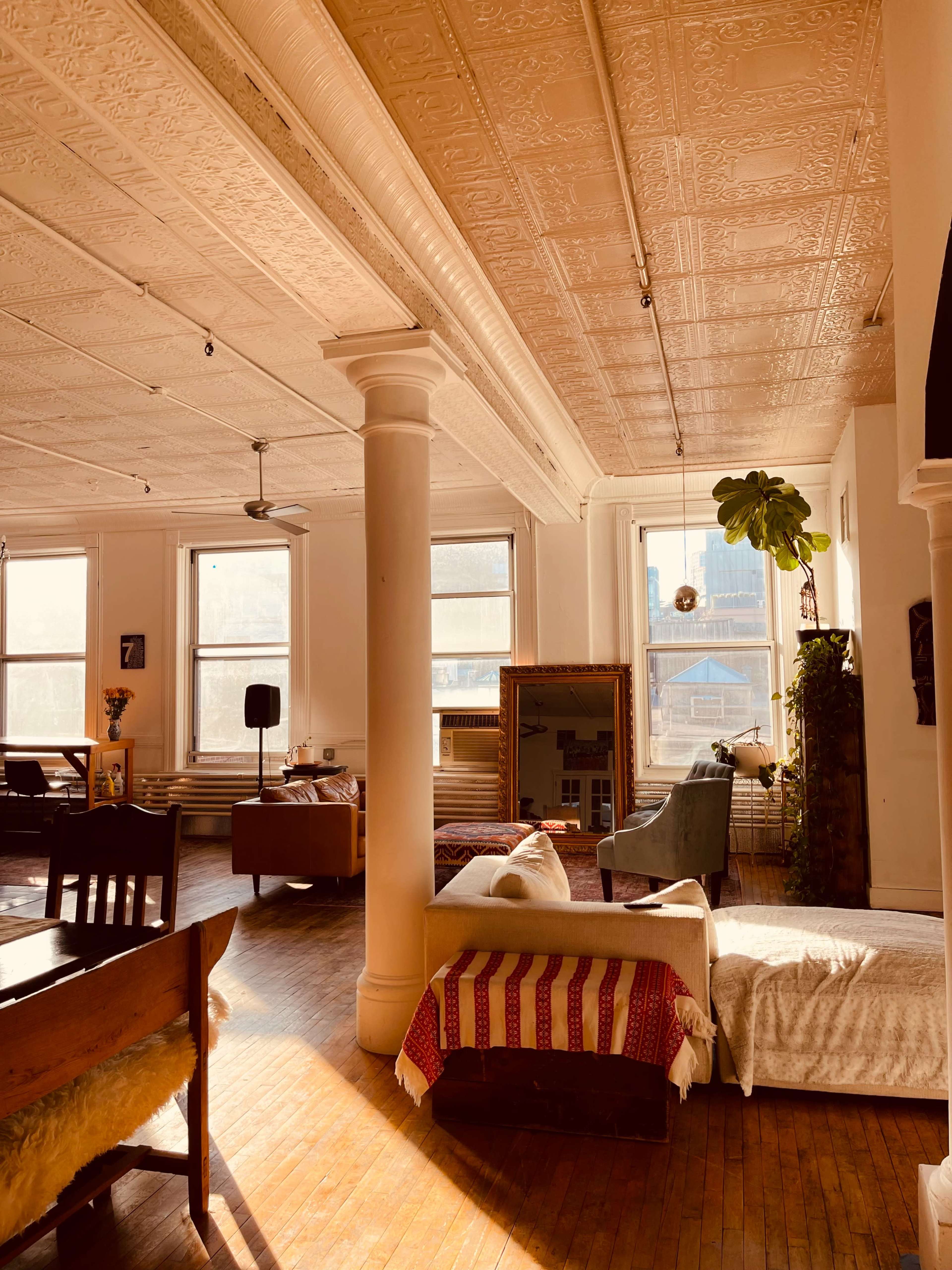 The image shows a spacious, well-lit room with large windows, wooden floors, and a mix of furniture, including a sofa and chairs.