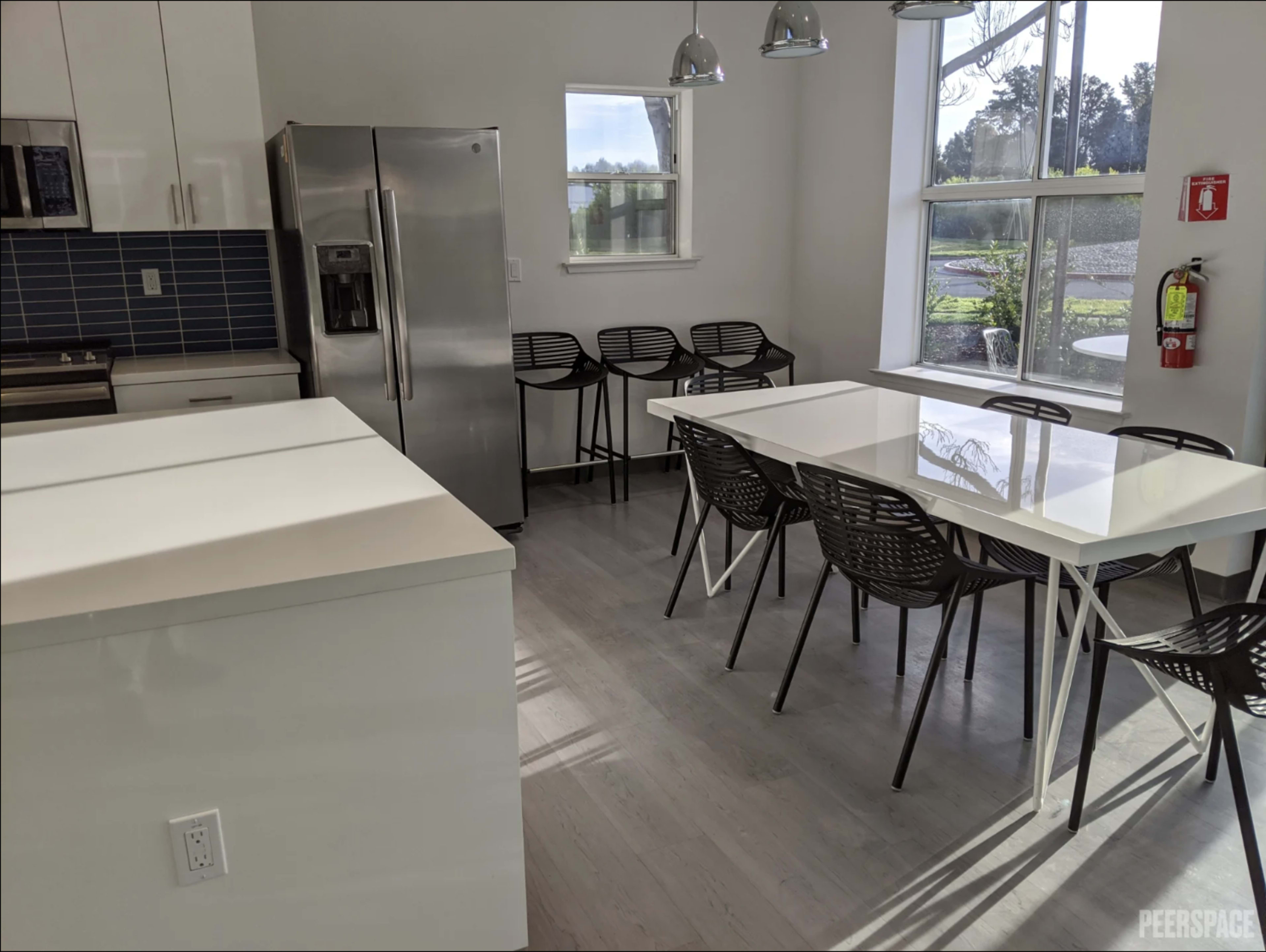 The image shows a modern kitchen and dining area with a stainless steel refrigerator, a long white table surrounded by black chairs, and large windows letting in natural light.