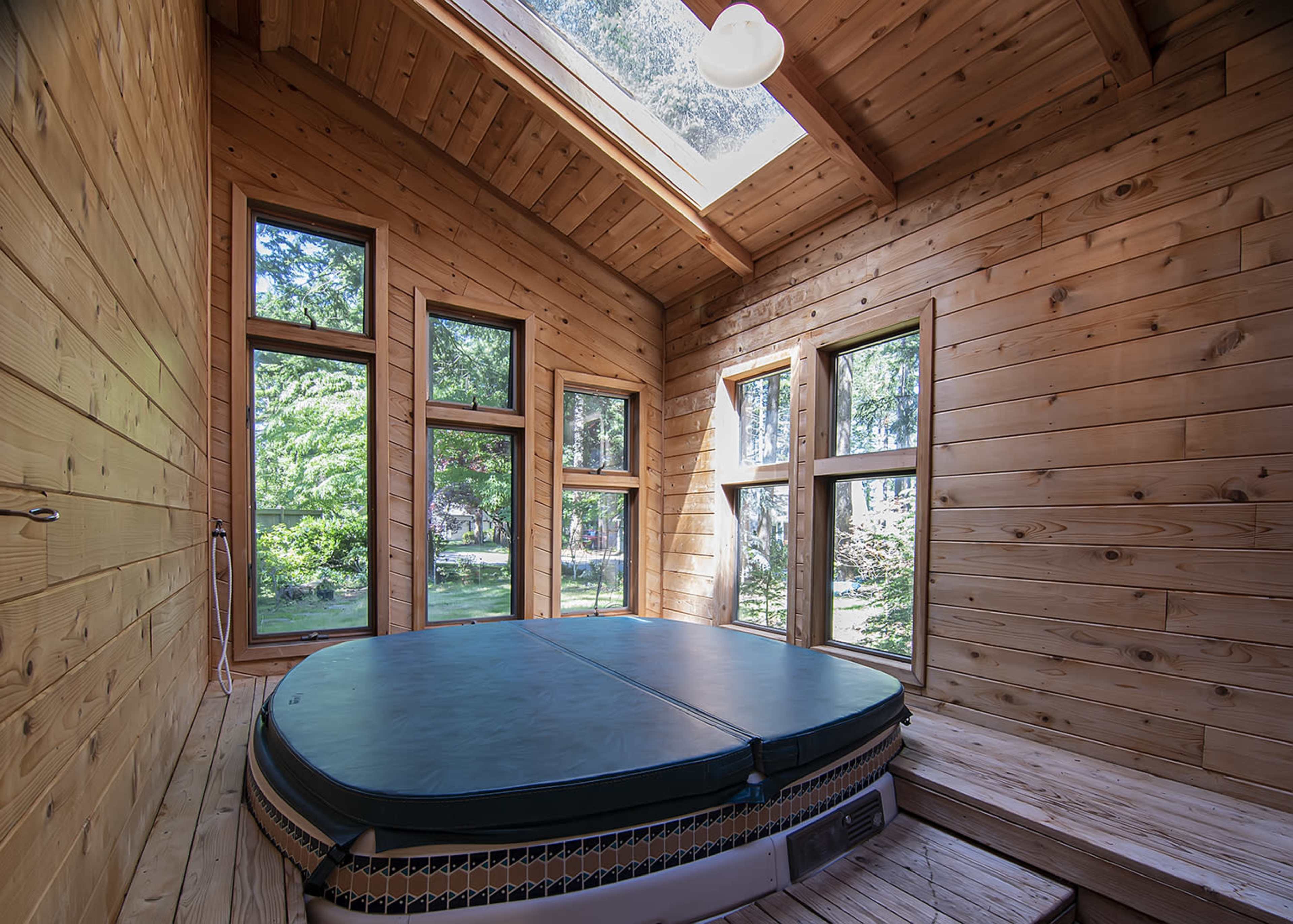 A wooden room features a circular hot tub surrounded by large windows that overlook greenery outside.
