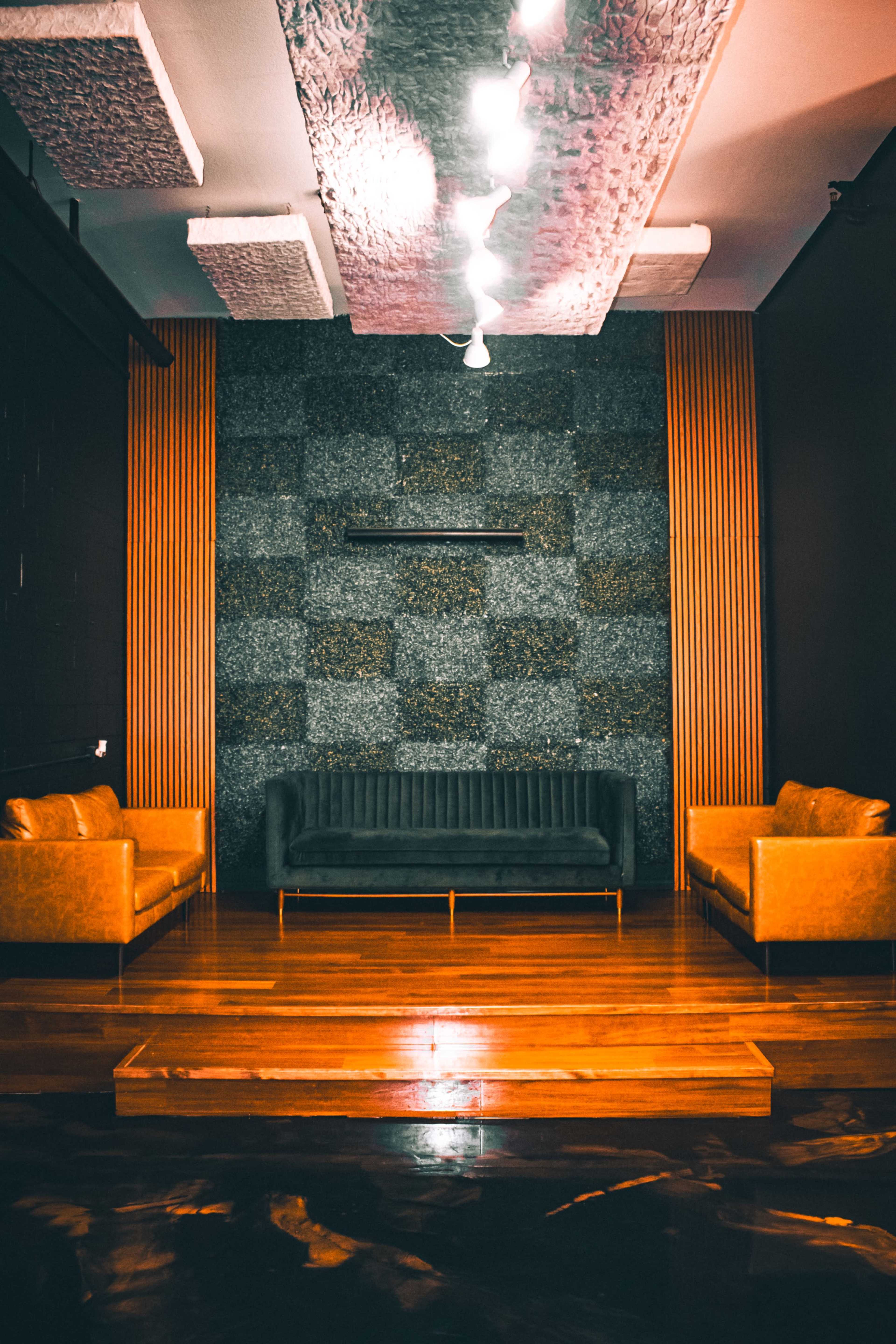 The image shows a stylish lounge area with a dark textured wall, wooden accents, and a pair of brown sofas facing a central wooden platform.