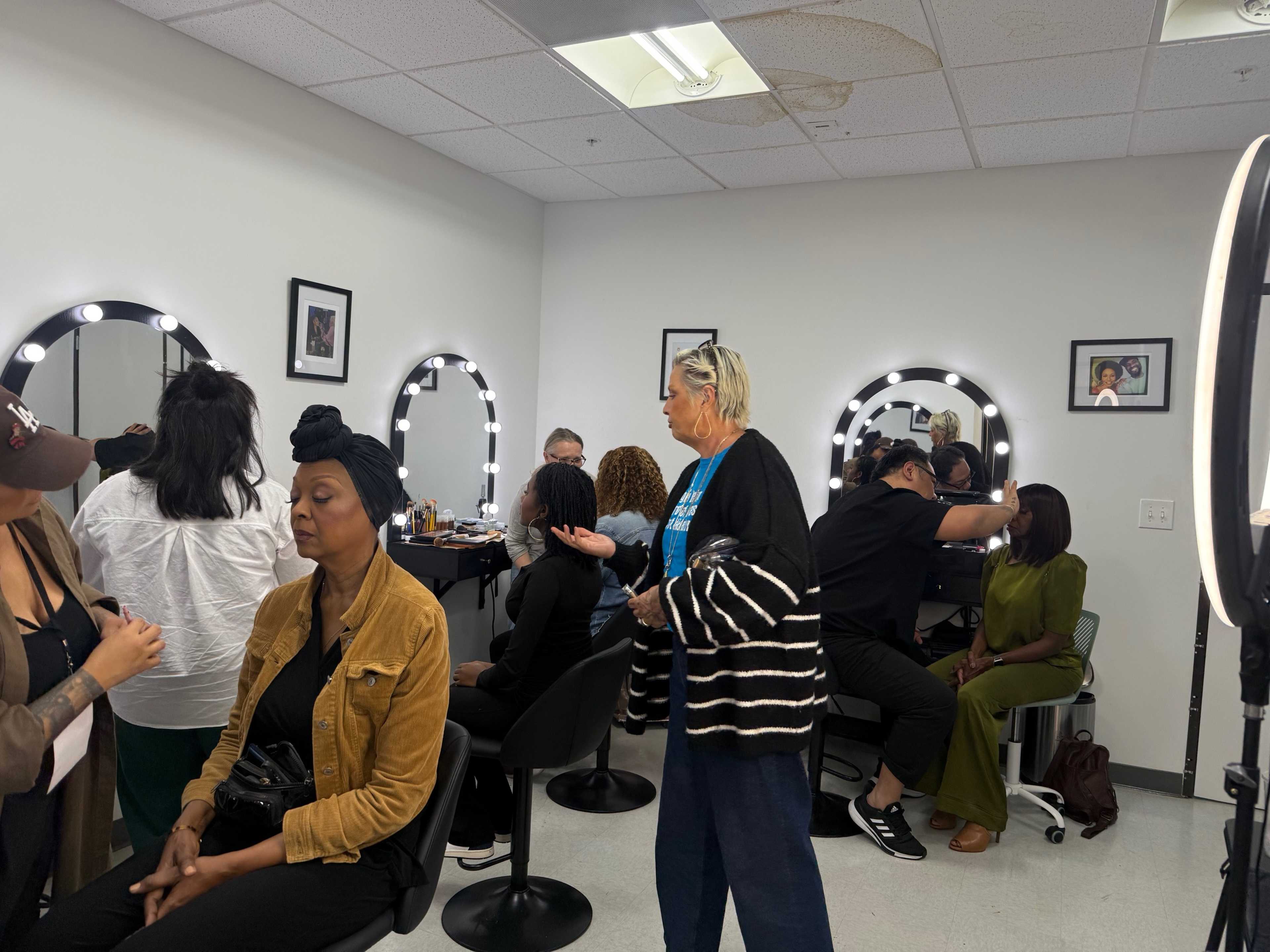 A makeup studio is filled with several people receiving makeup applications, while others are engaged in conversation and preparation.
