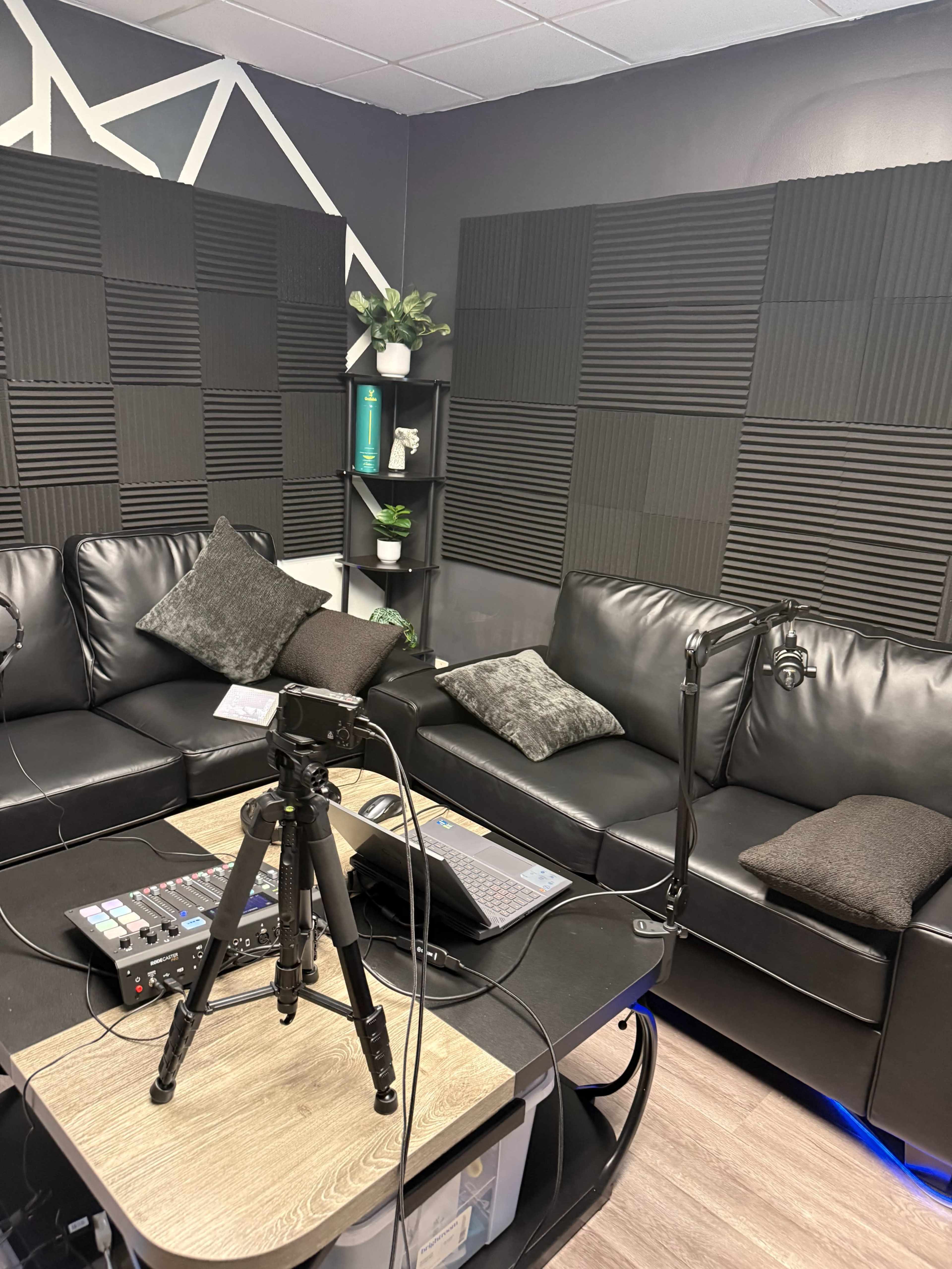 The image shows a recording space featuring a black couch with decorative pillows, a laptop, a microphone on a stand, and soundproof wall panels.
