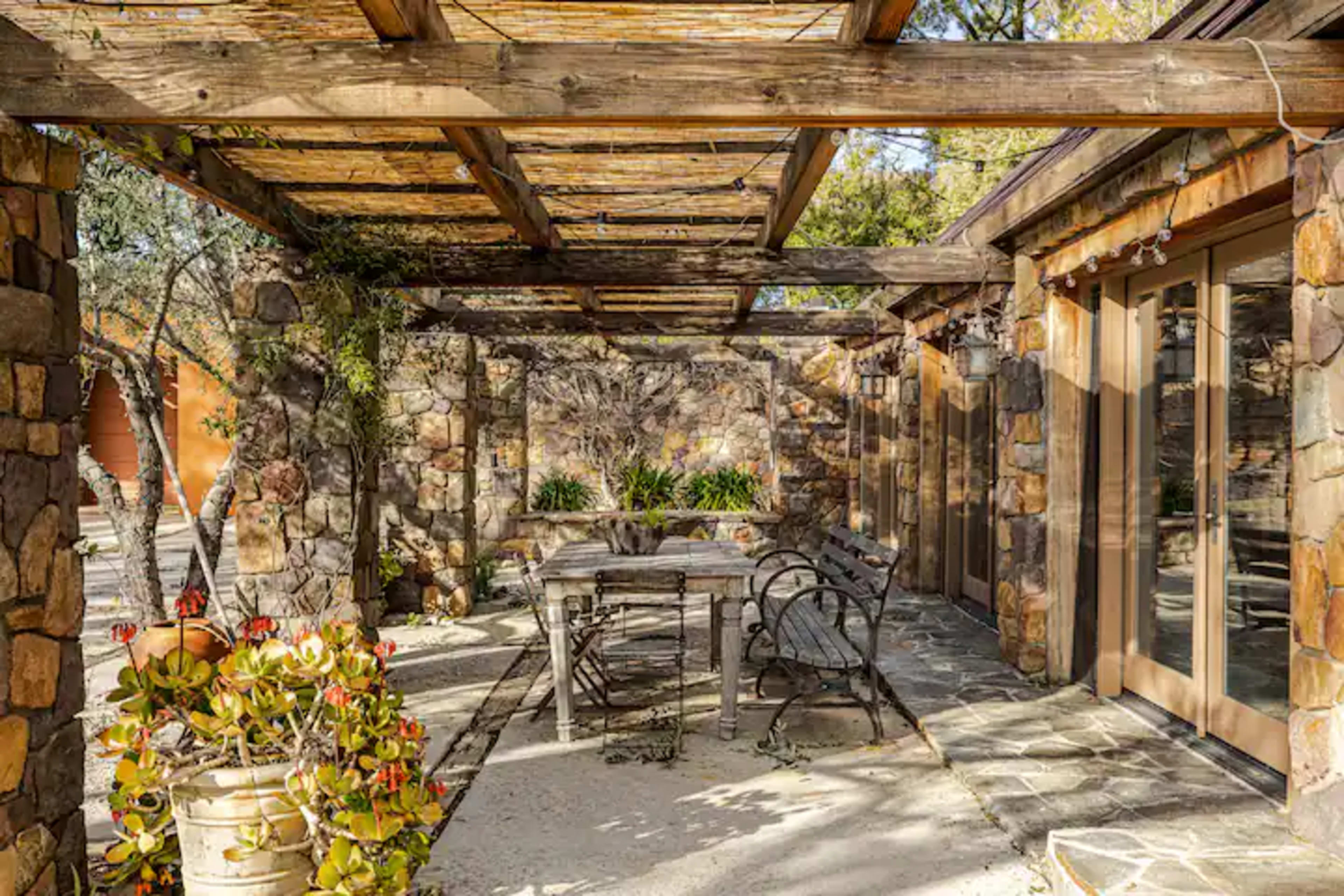 The image shows a stone patio area with a wooden pergola, featuring a dining table, chairs, and potted plants.