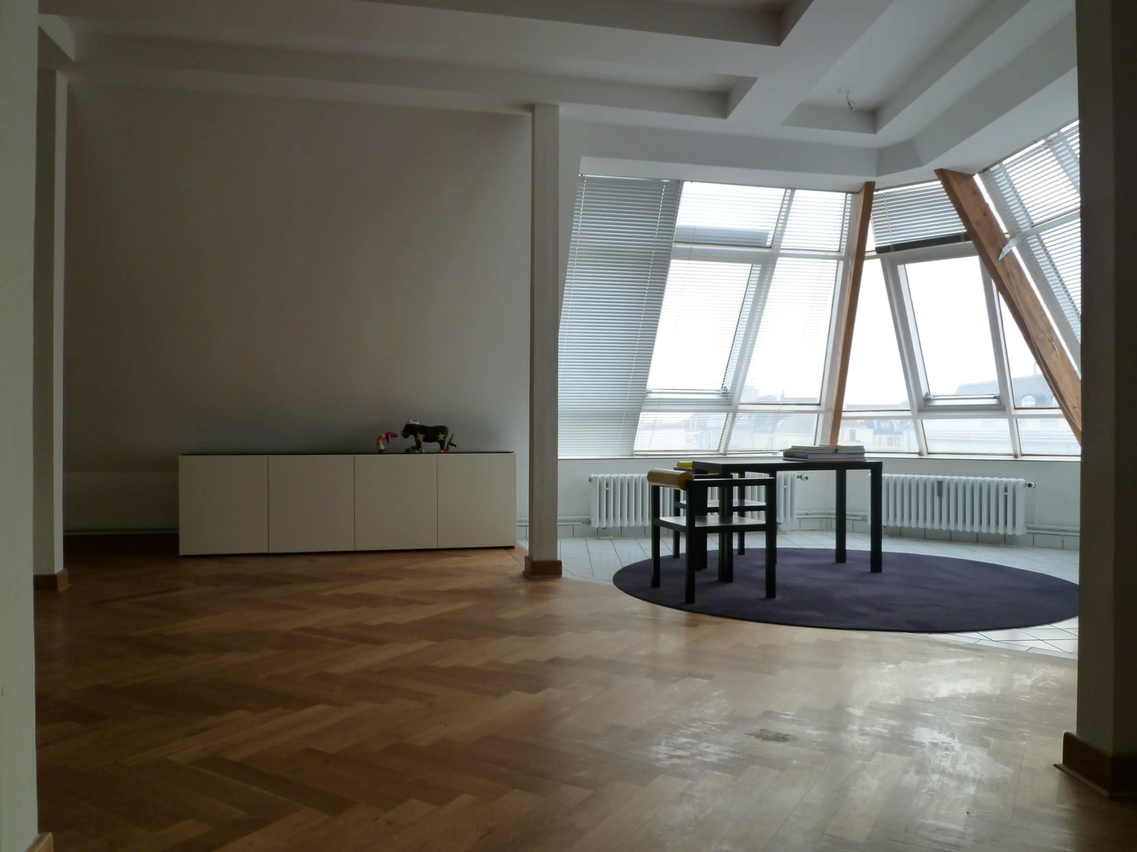 A spacious room features a table and chairs near large windows with an angular design, set on a hardwood floor.