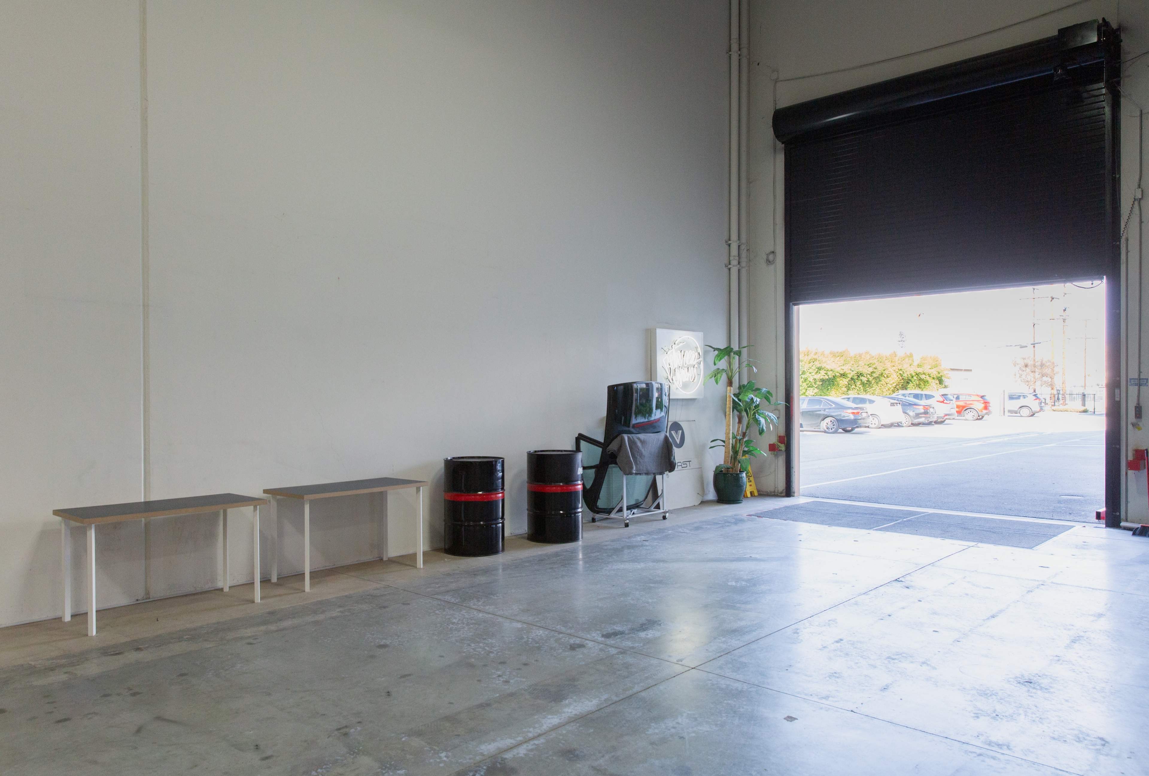 An empty warehouse space with two tables, two black barrels, a chair, a potted plant, and a large open roll-up door leading to an exterior parking area.