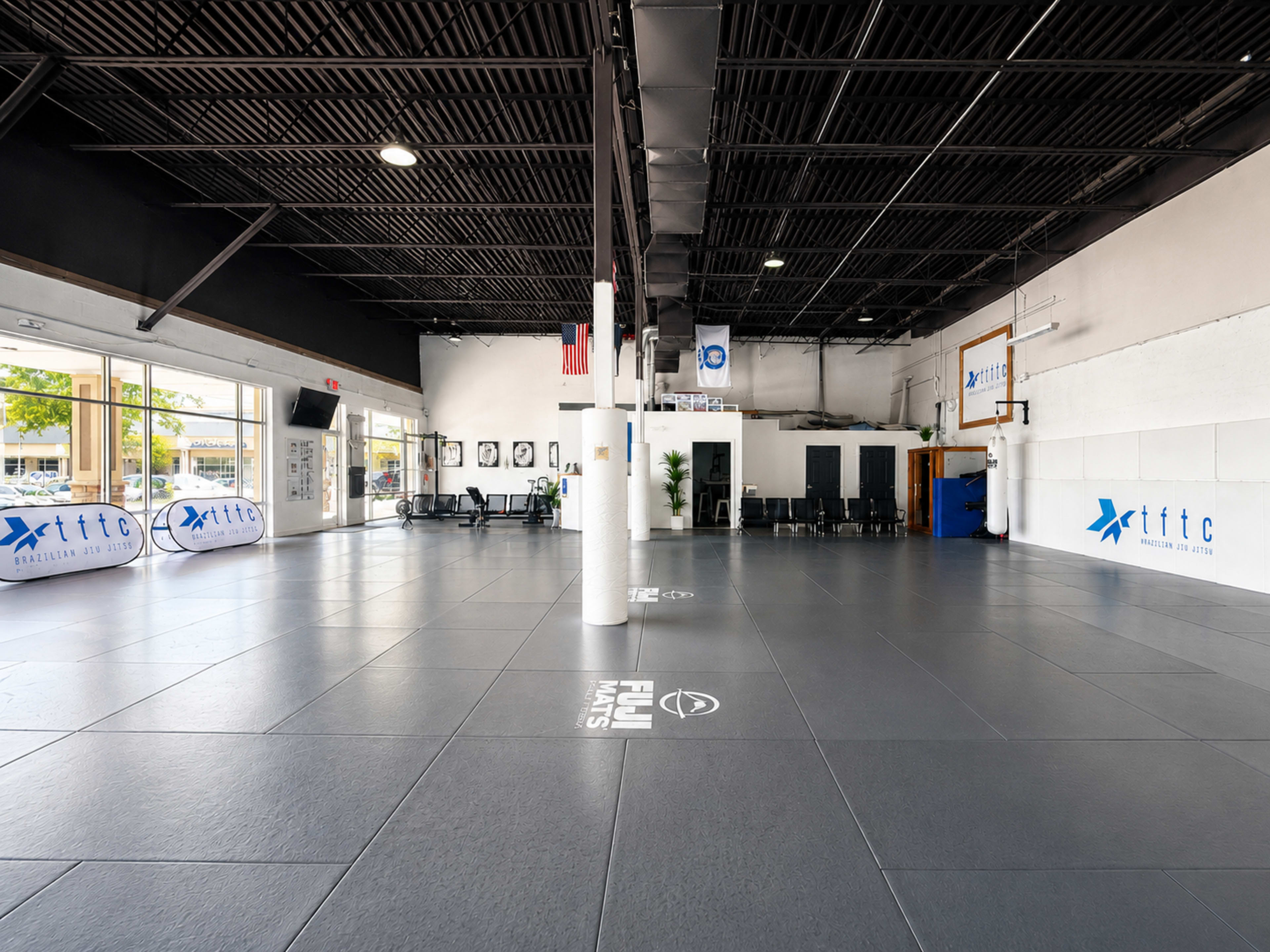 The image shows a spacious training facility with dark mats, padded walls, and various equipment arranged along the sides.